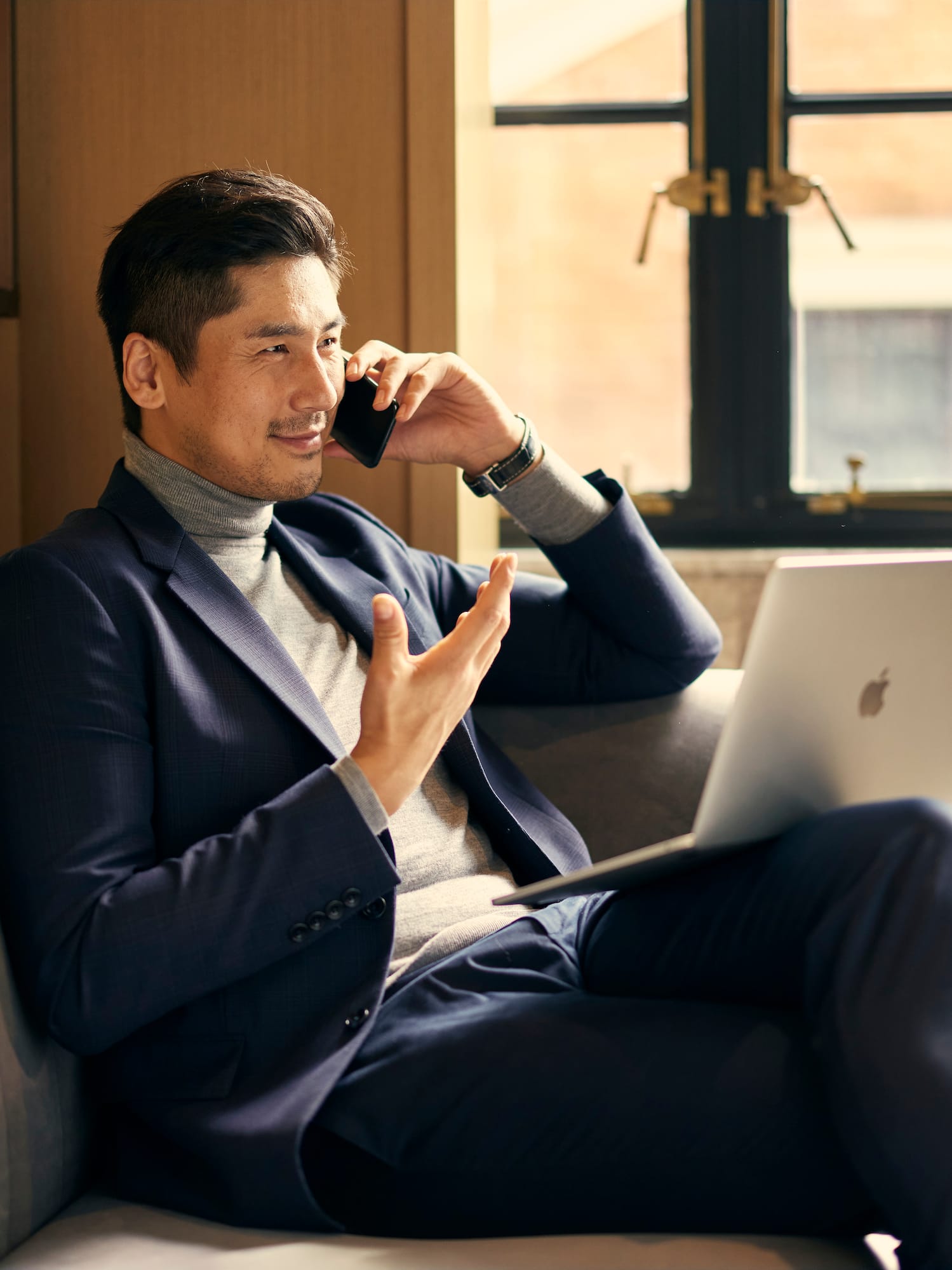 a man sitting on a couch talking on a phone and using a laptop