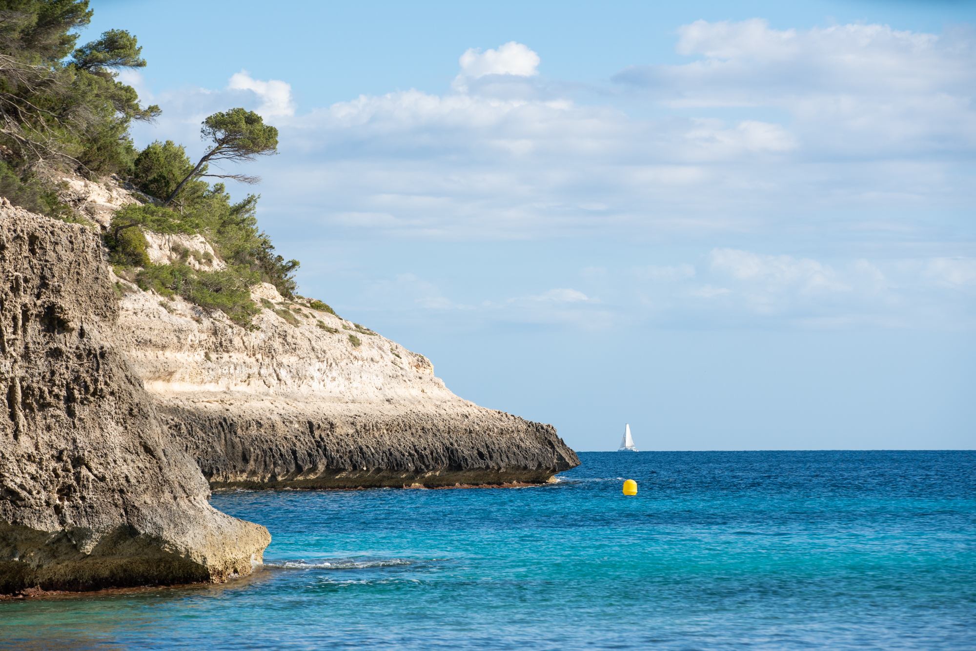 a rocky cliff with trees and a sailboat in the water