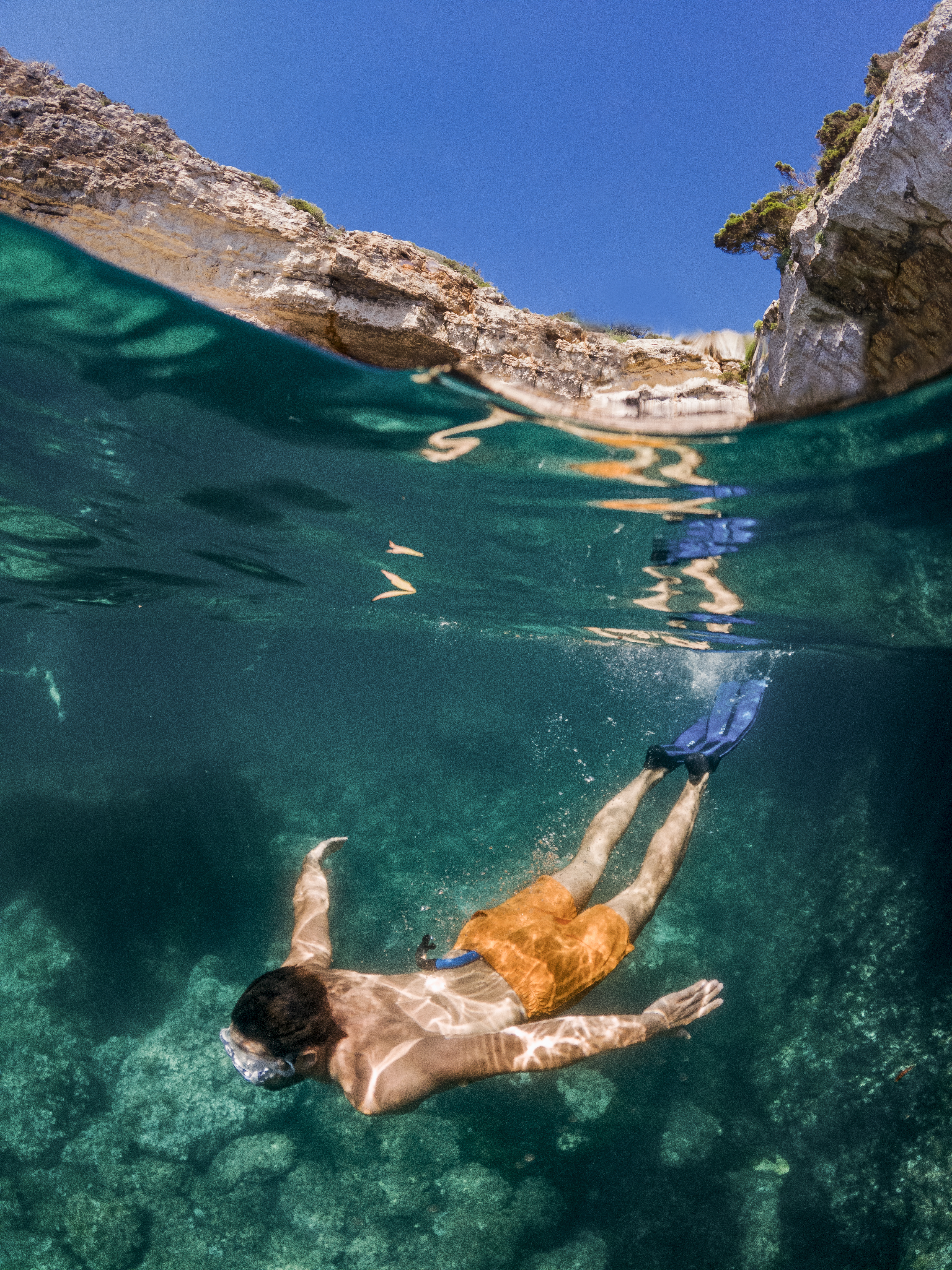 Man snorkeling in turquoise water below a sunny rocky cliff.