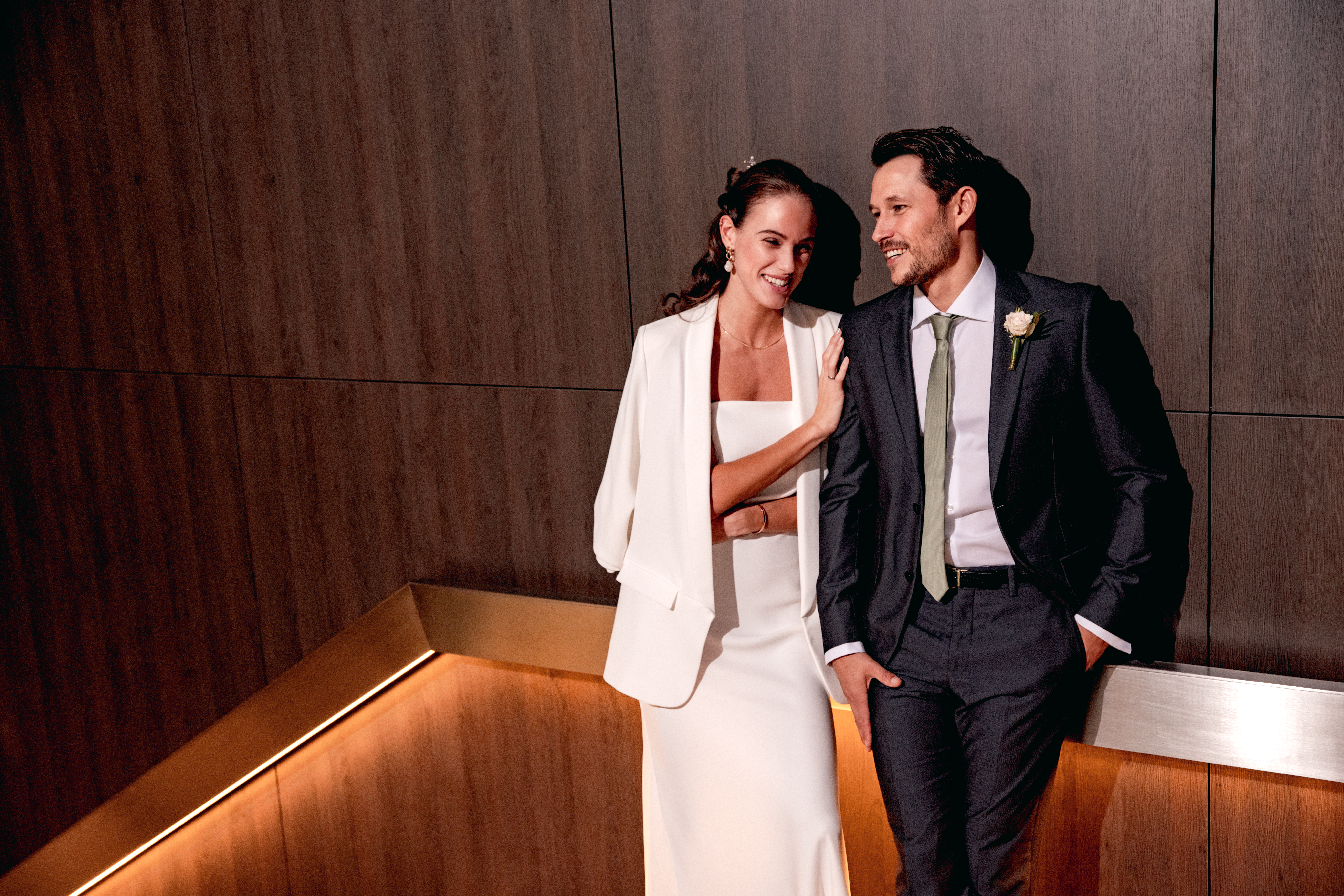 Bride and groom smiling, elegantly dressed, against dark wood interior.