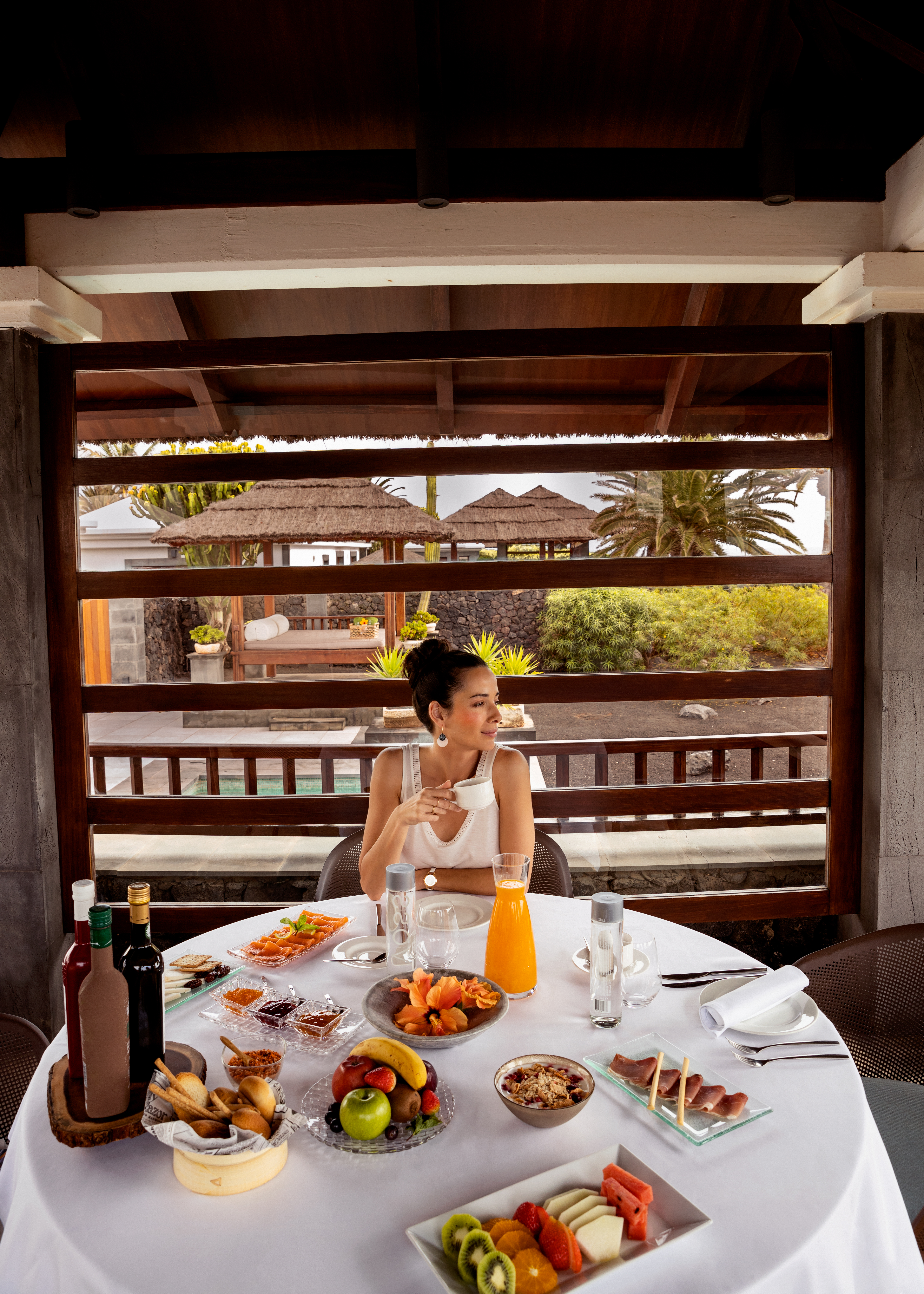 Woman enjoying lavish outdoor breakfast with coffee, fruits, and tropical view.