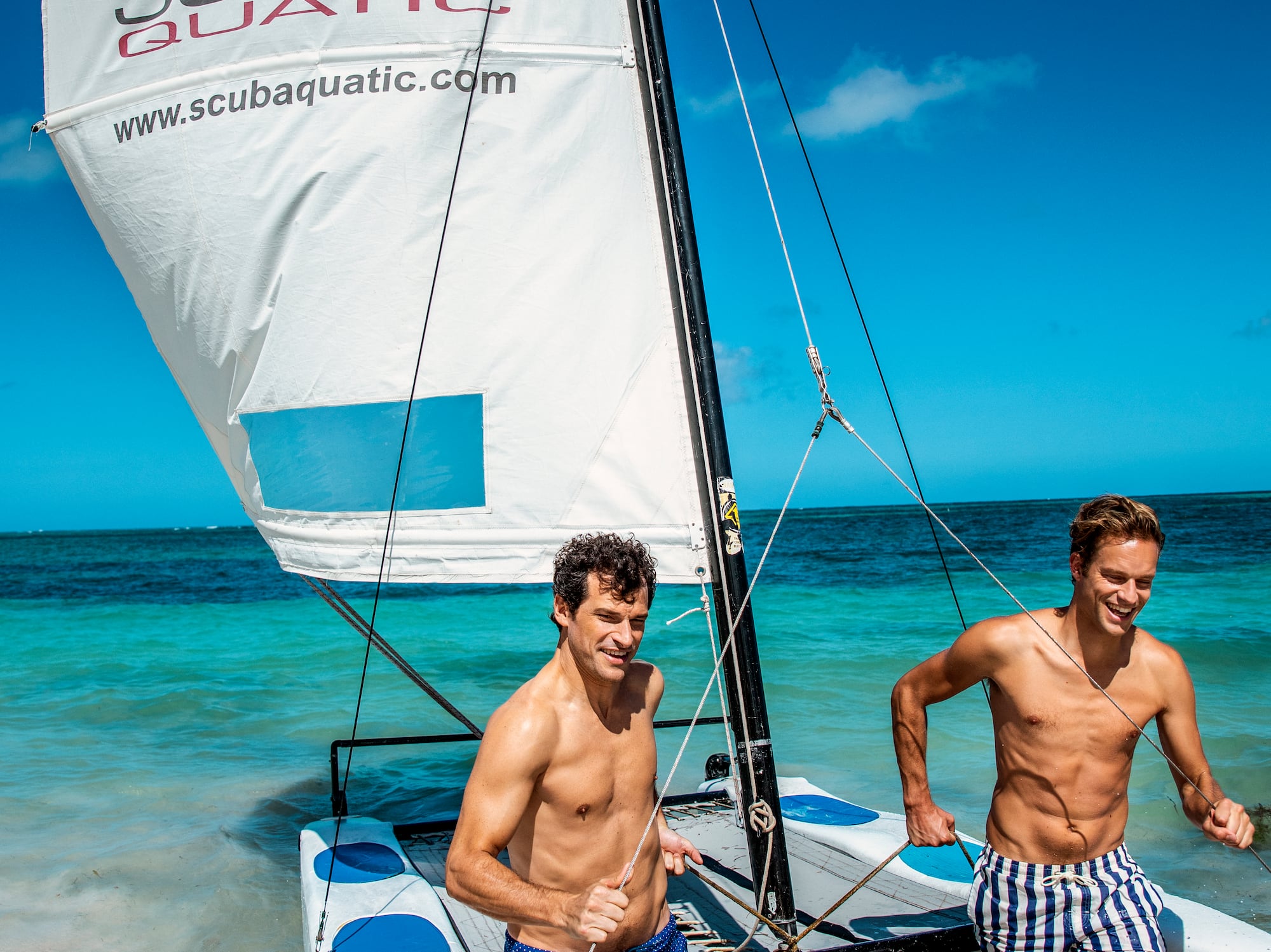 two men in swim trunks on a sailboat