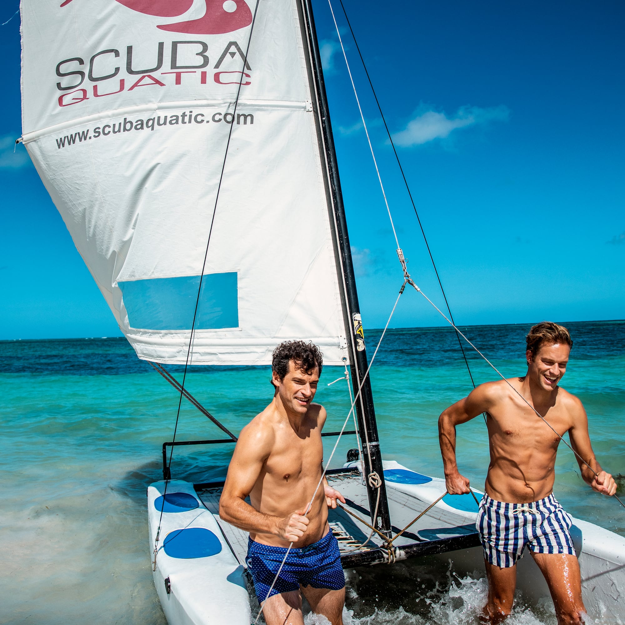two men in swim trunks on a sailboat