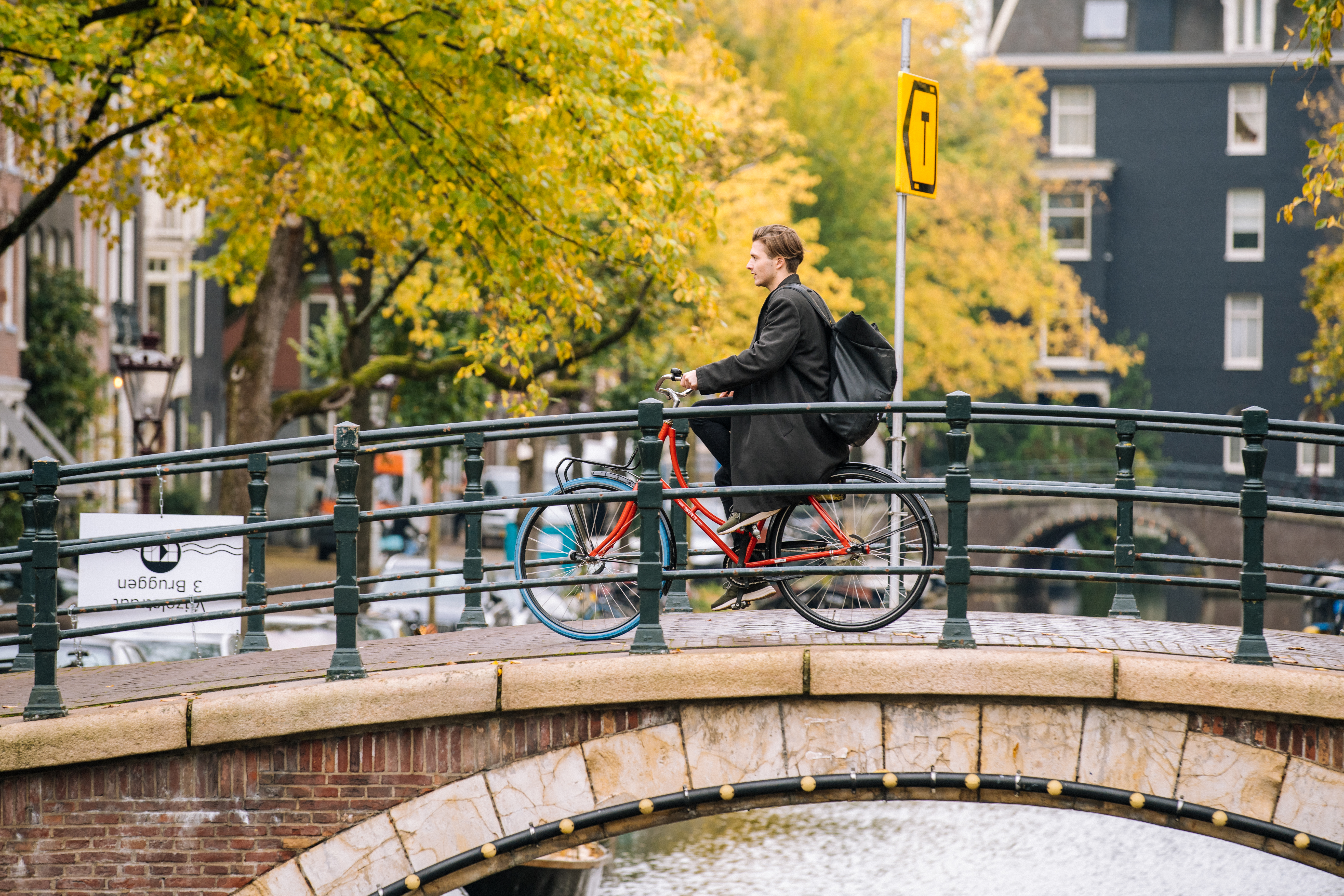 Man cycling red bike over Amsterdam canal bridge, autumn leaves.