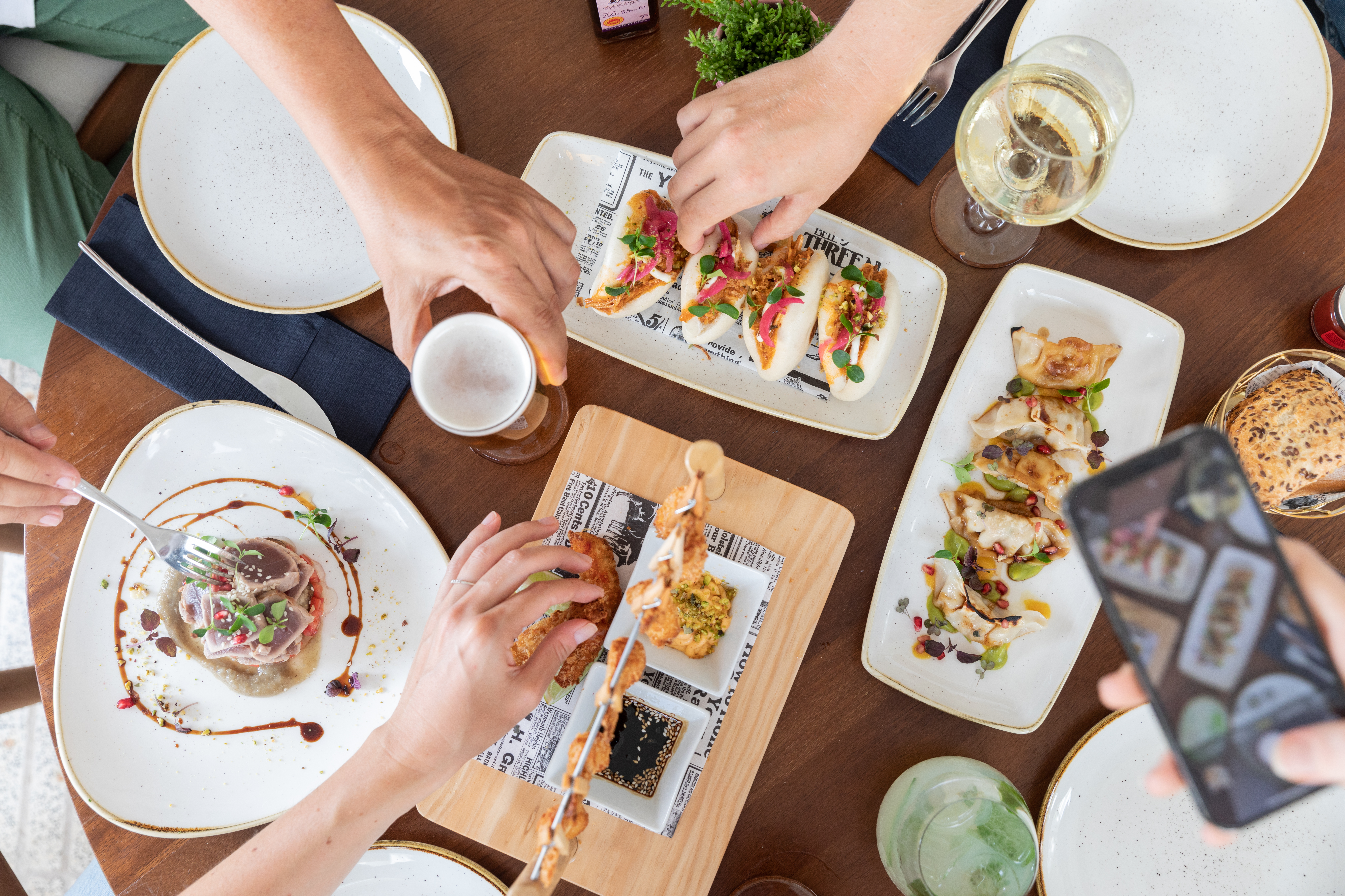 Hands sharing gourmet restaurant appetizers: bao buns, dumplings, tuna, and drinks.