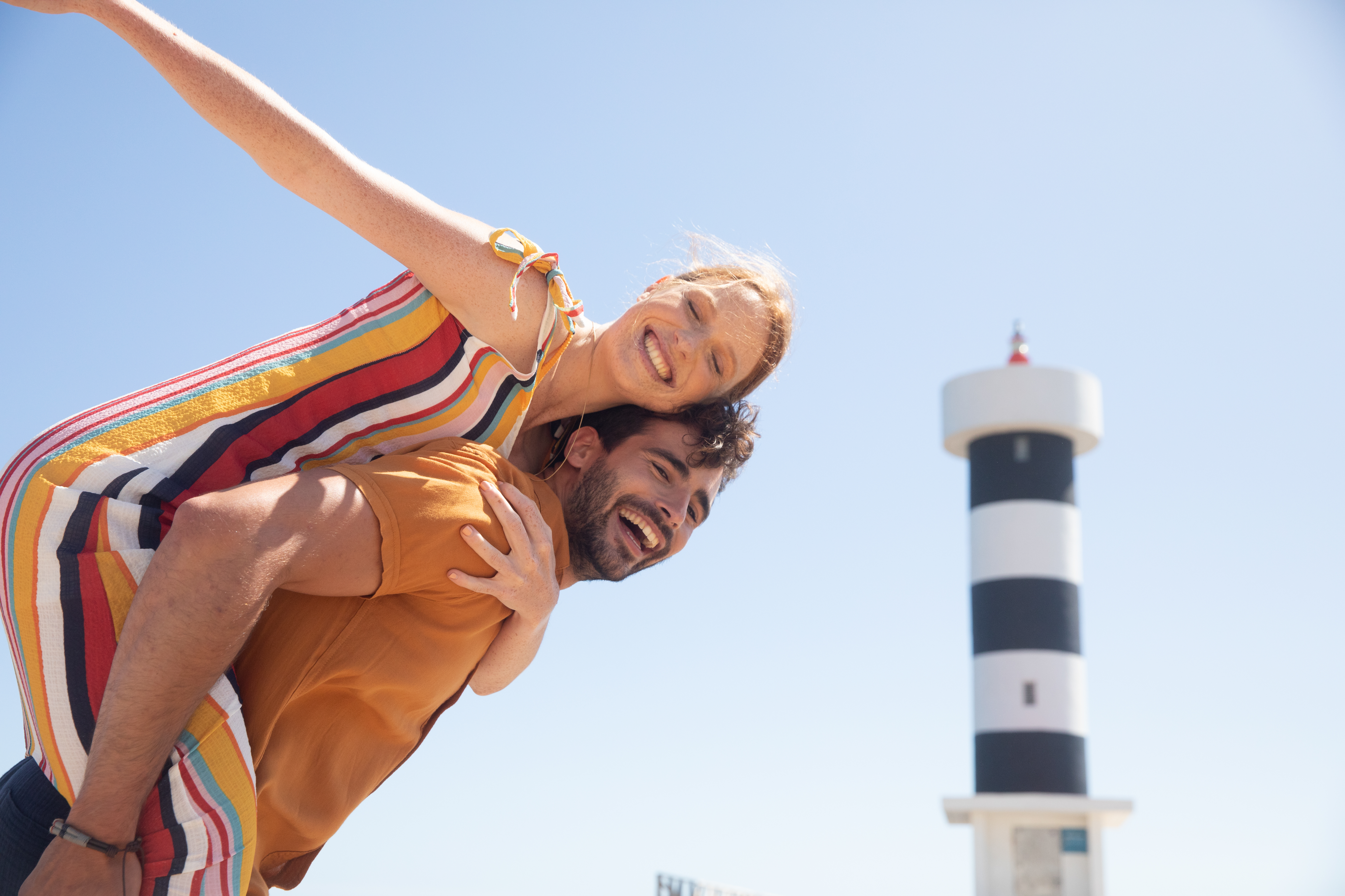 Smiling couple on piggyback ride, lighthouse in background, sunny day.
