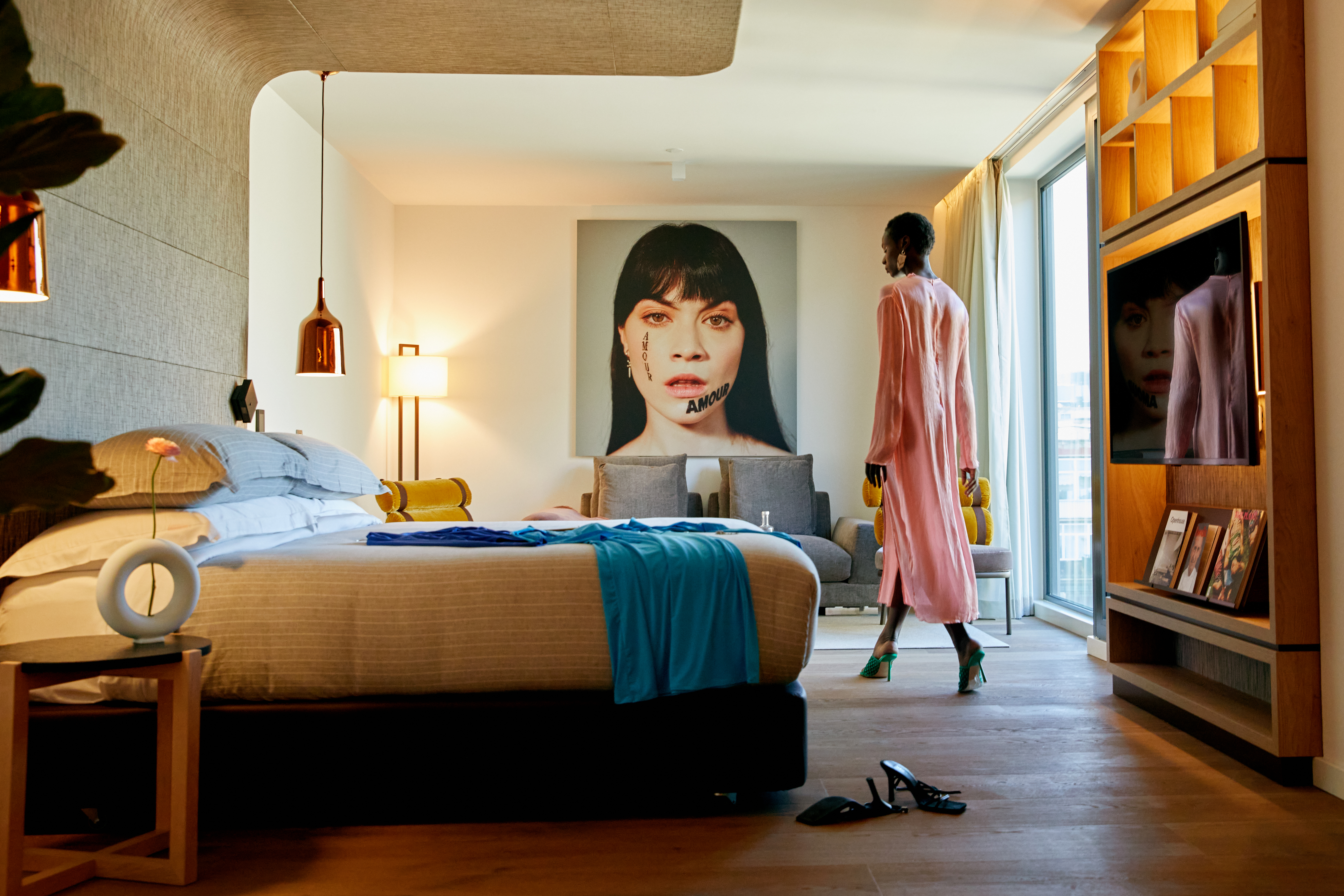 Contemporary hotel bedroom with large bed, striking art, and woman by window.