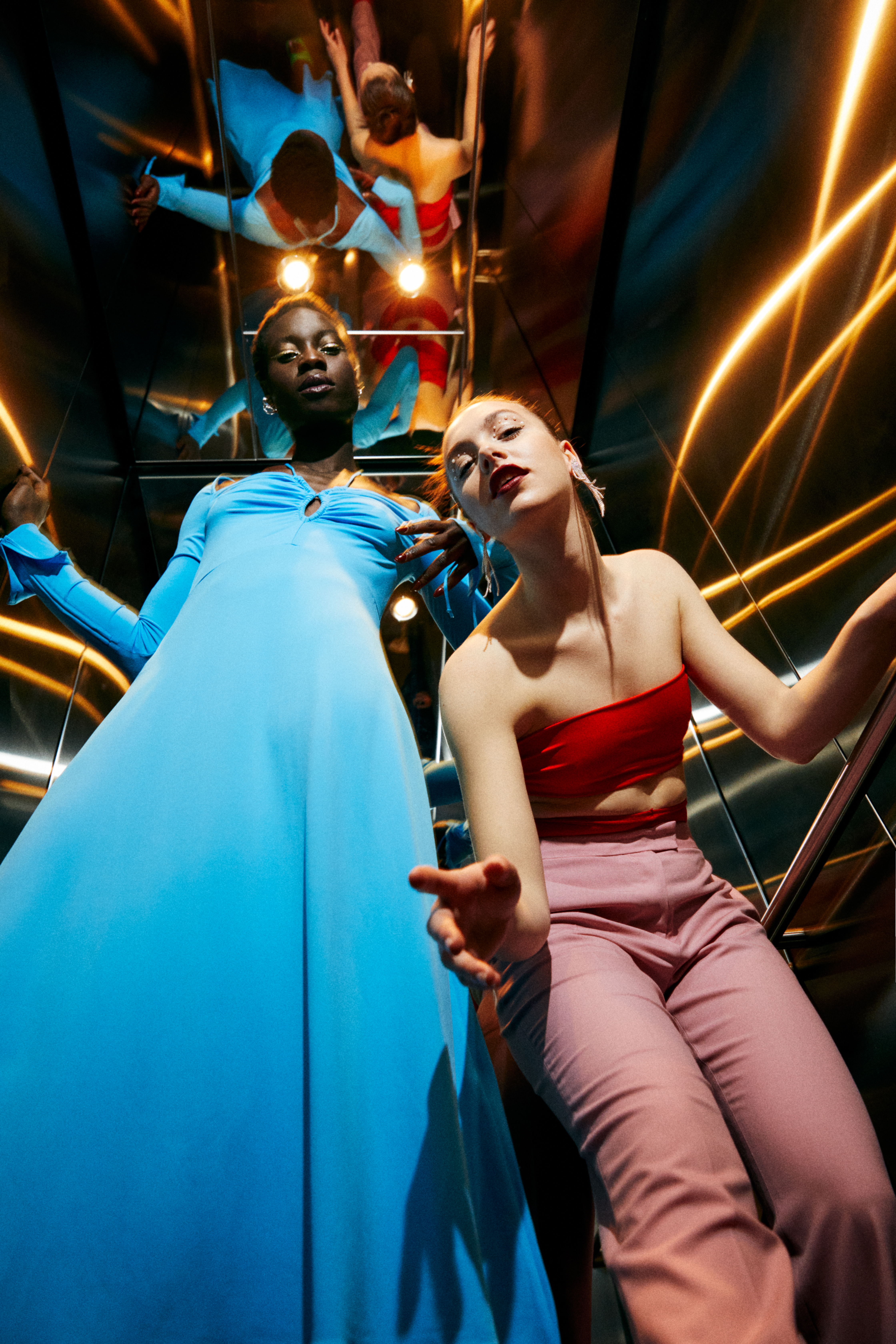Two women in blue dress and red top, mirrored elevator with golden lights.
