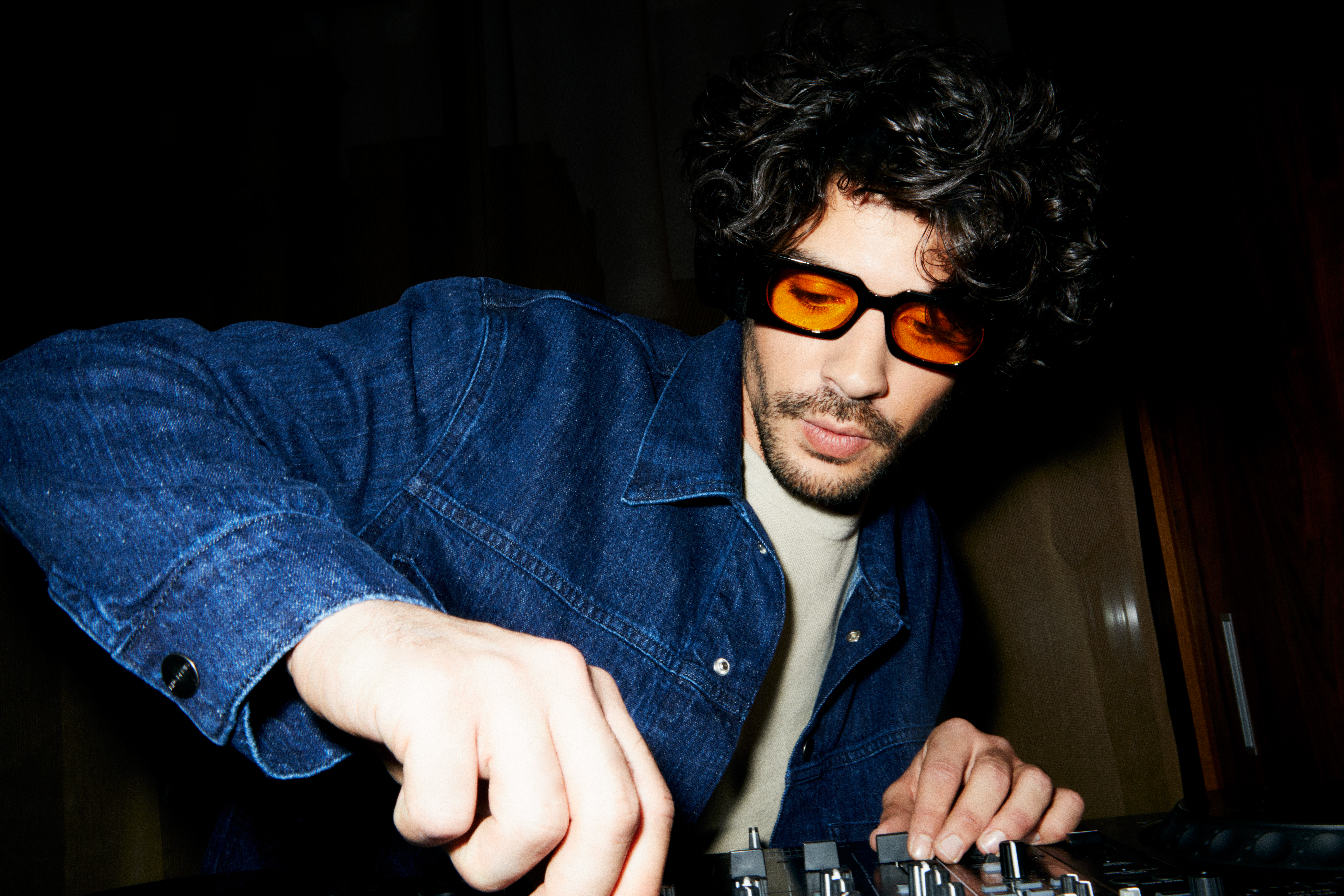 DJ in denim jacket, orange glasses, mixing music on a club mixer.