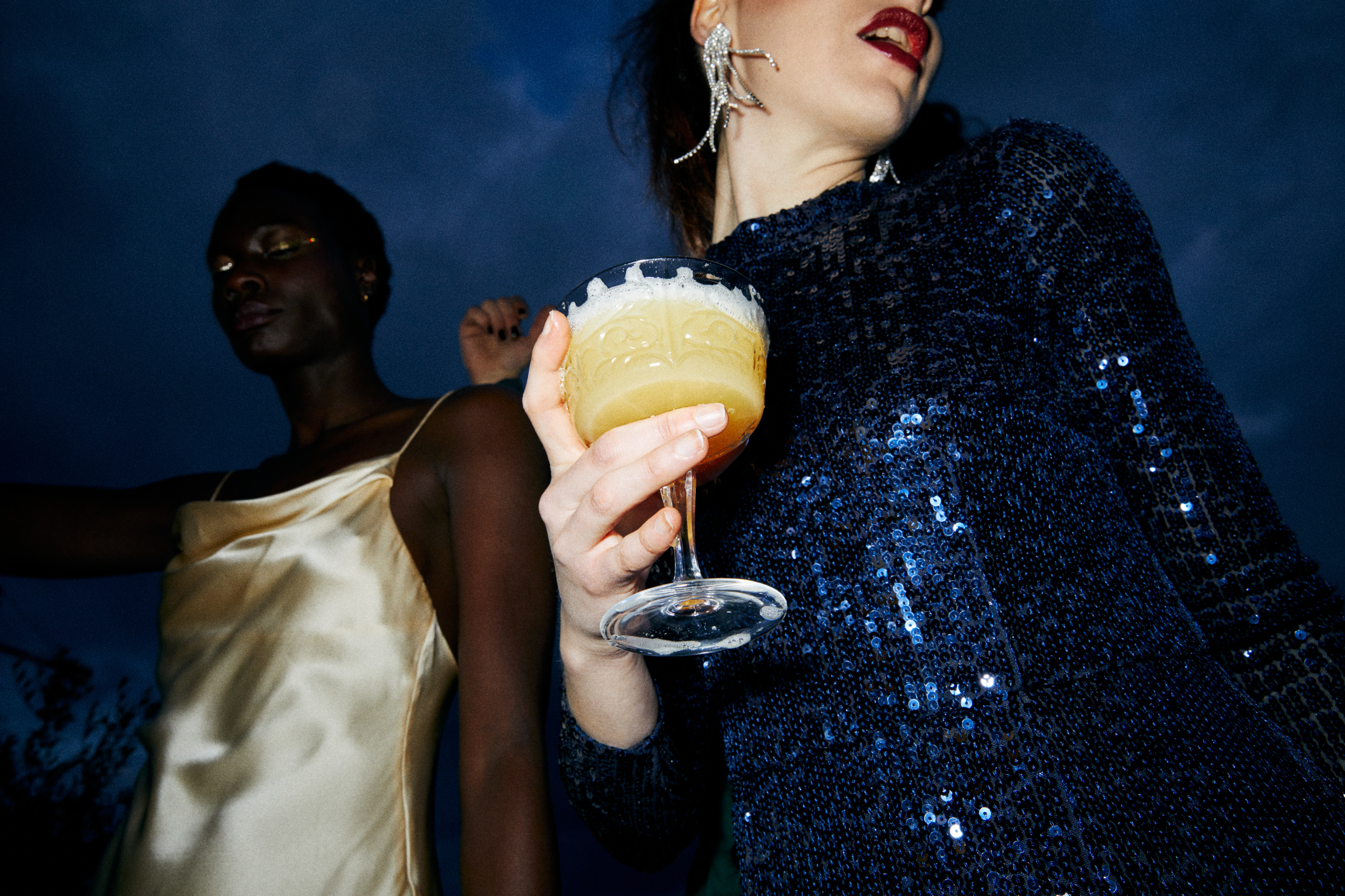 Woman in blue sequin top holding cocktail, friend dancing at party.
