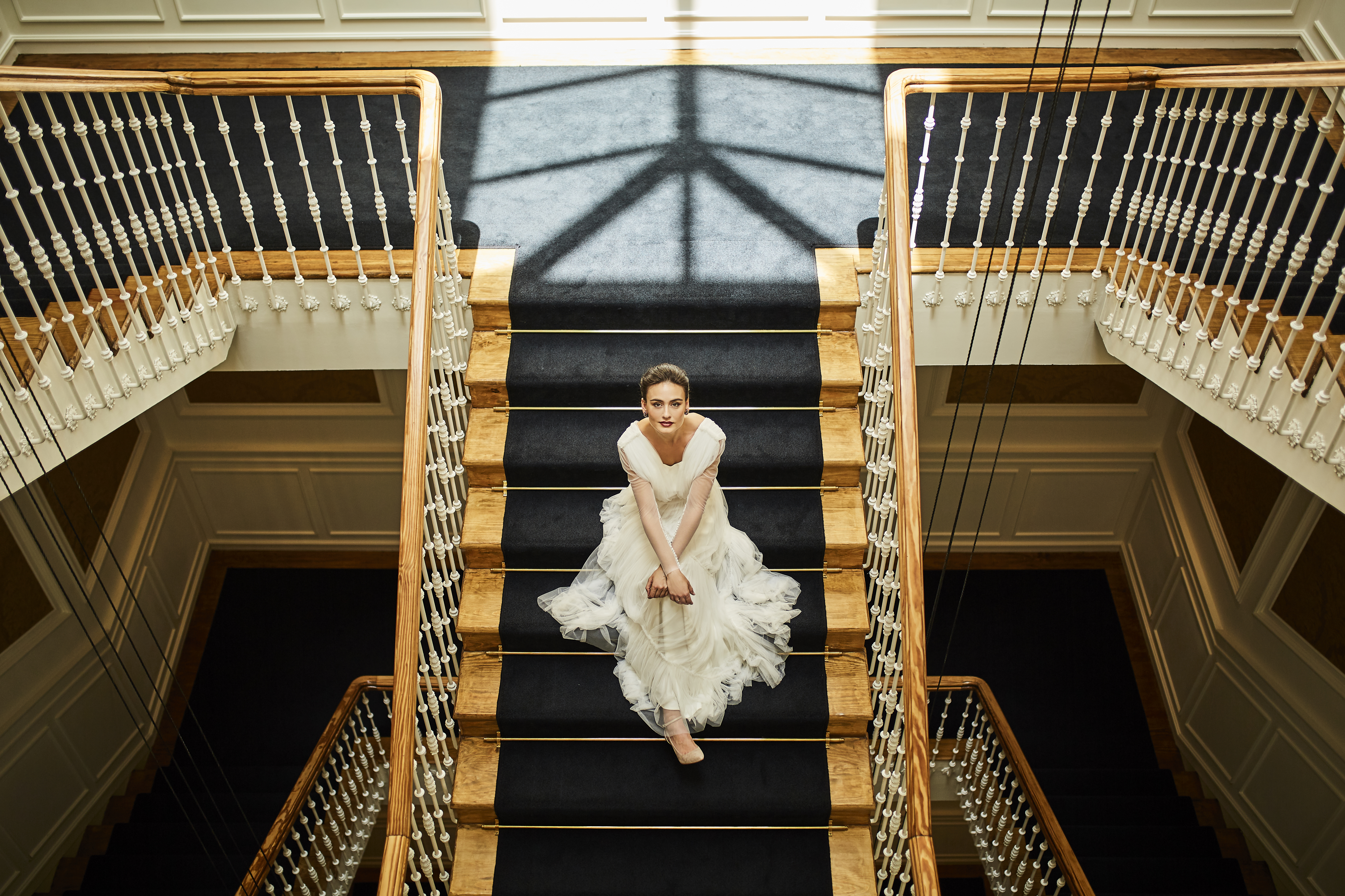 Bride in white wedding dress sitting on grand elegant staircase with dark carpet.