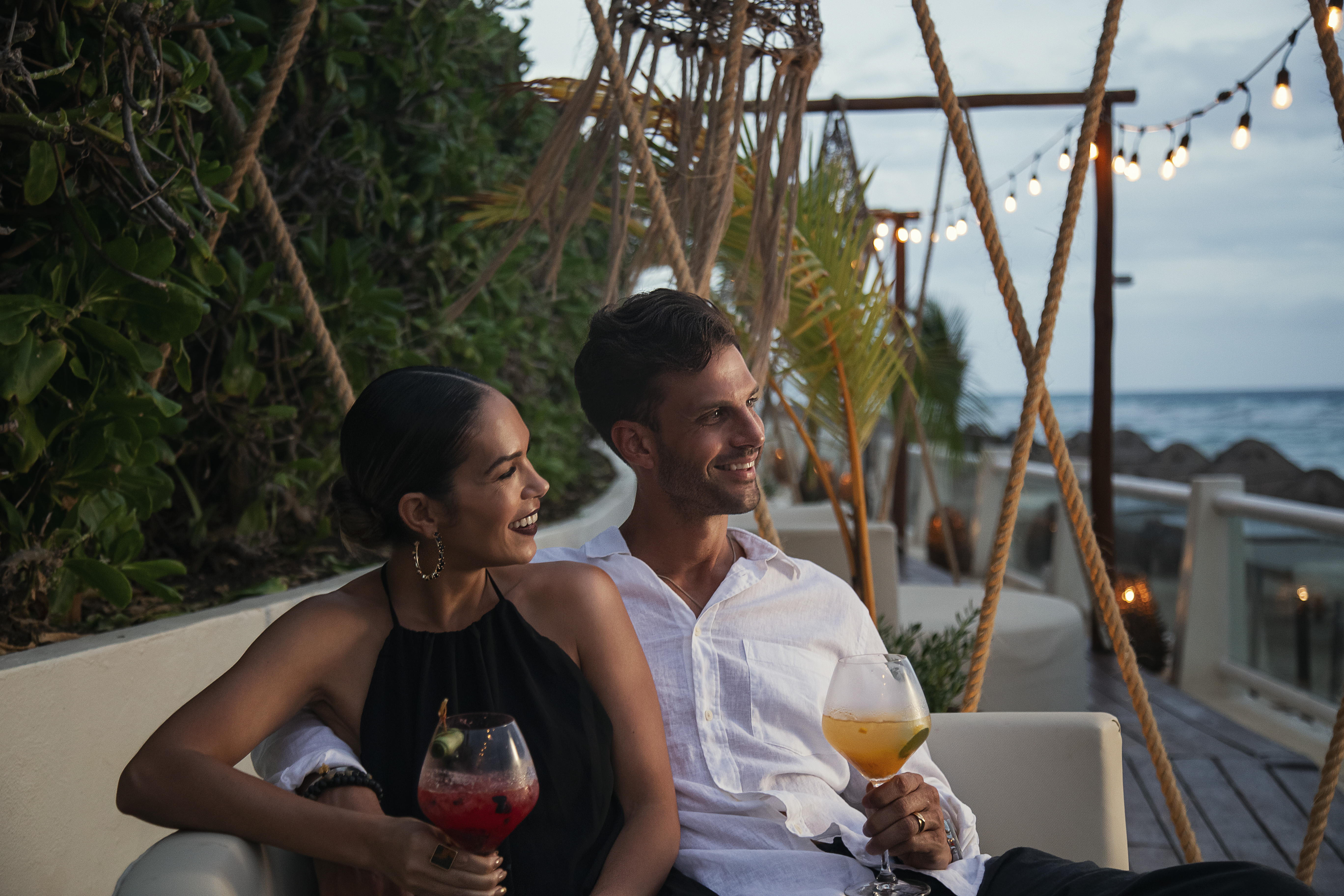 Smiling couple enjoying cocktails at a romantic beachside lounge with string lights.