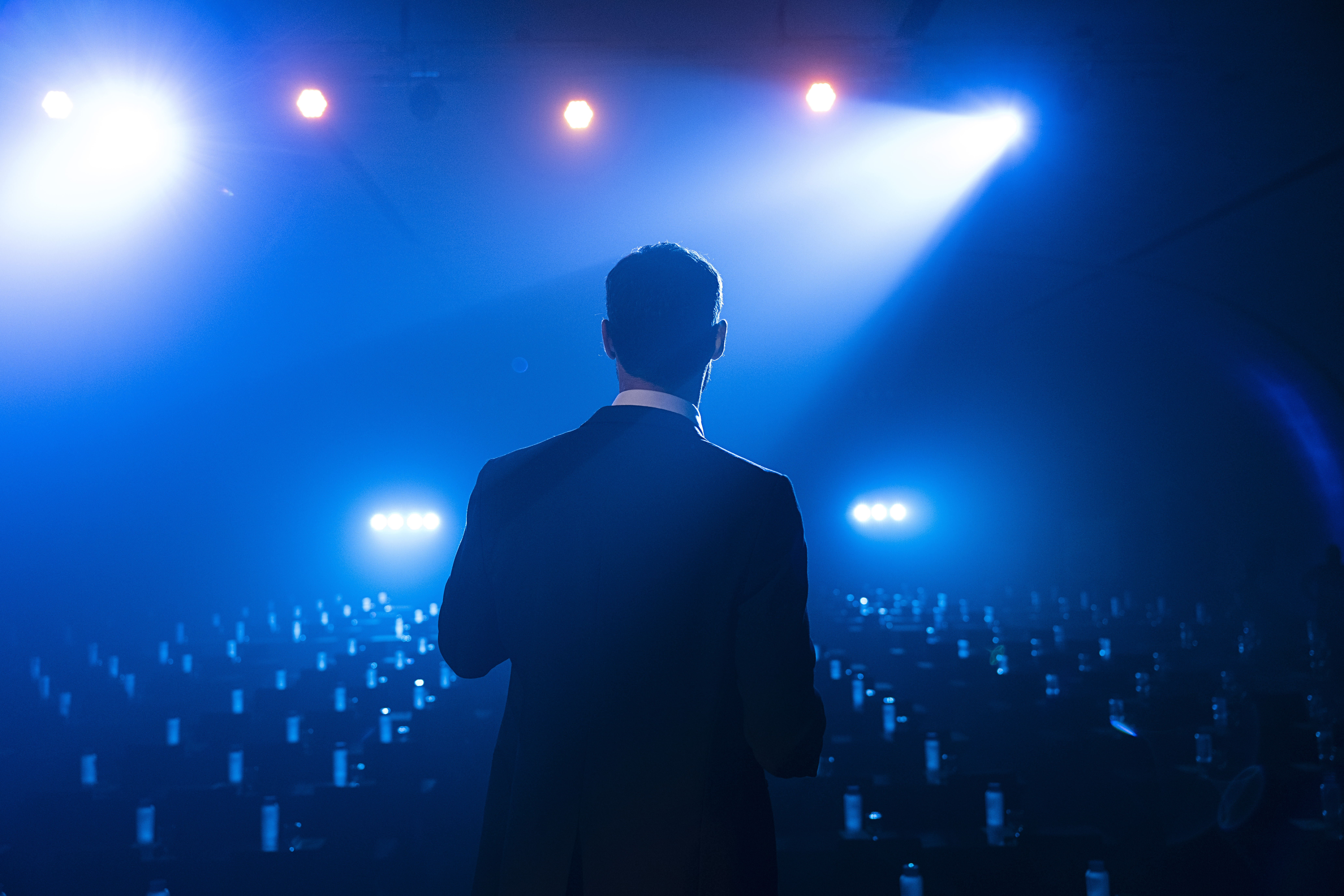 Presenter on stage under blue spotlights, facing a dark conference audience.