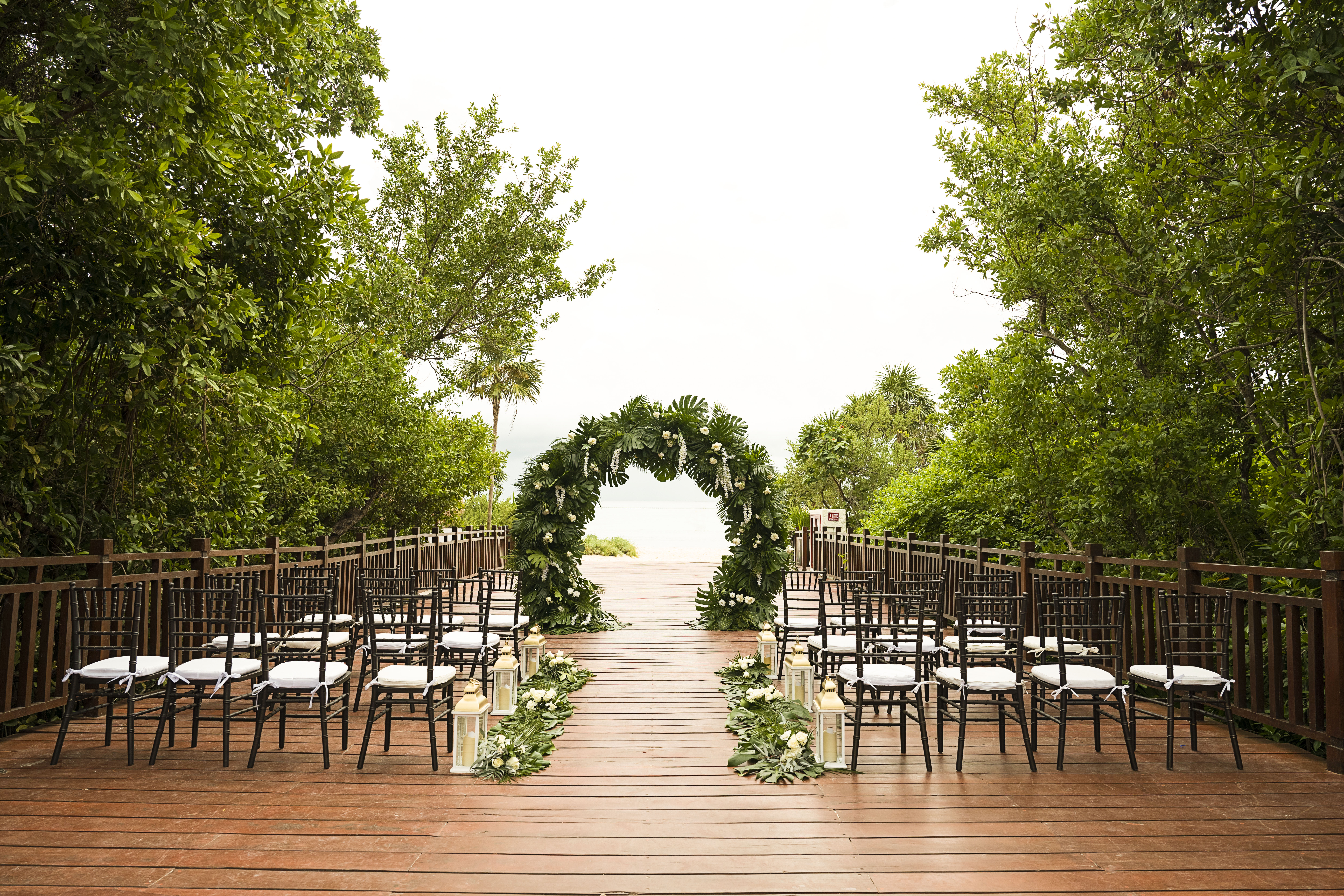 Beach wedding ceremony with tropical floral arch, guest chairs, wooden aisle.