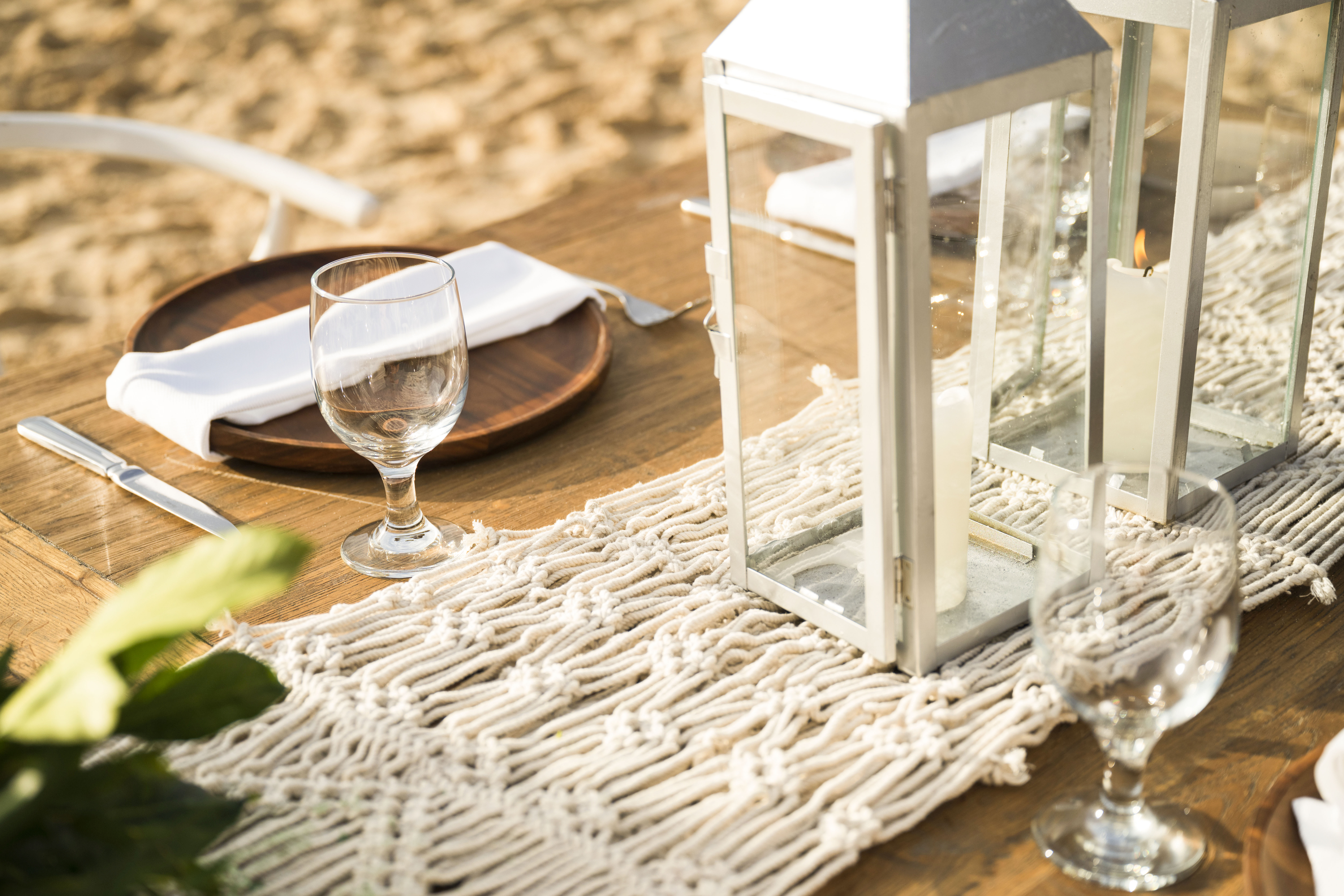Beach table setting with lit lantern, macrame runner, and wine glass.