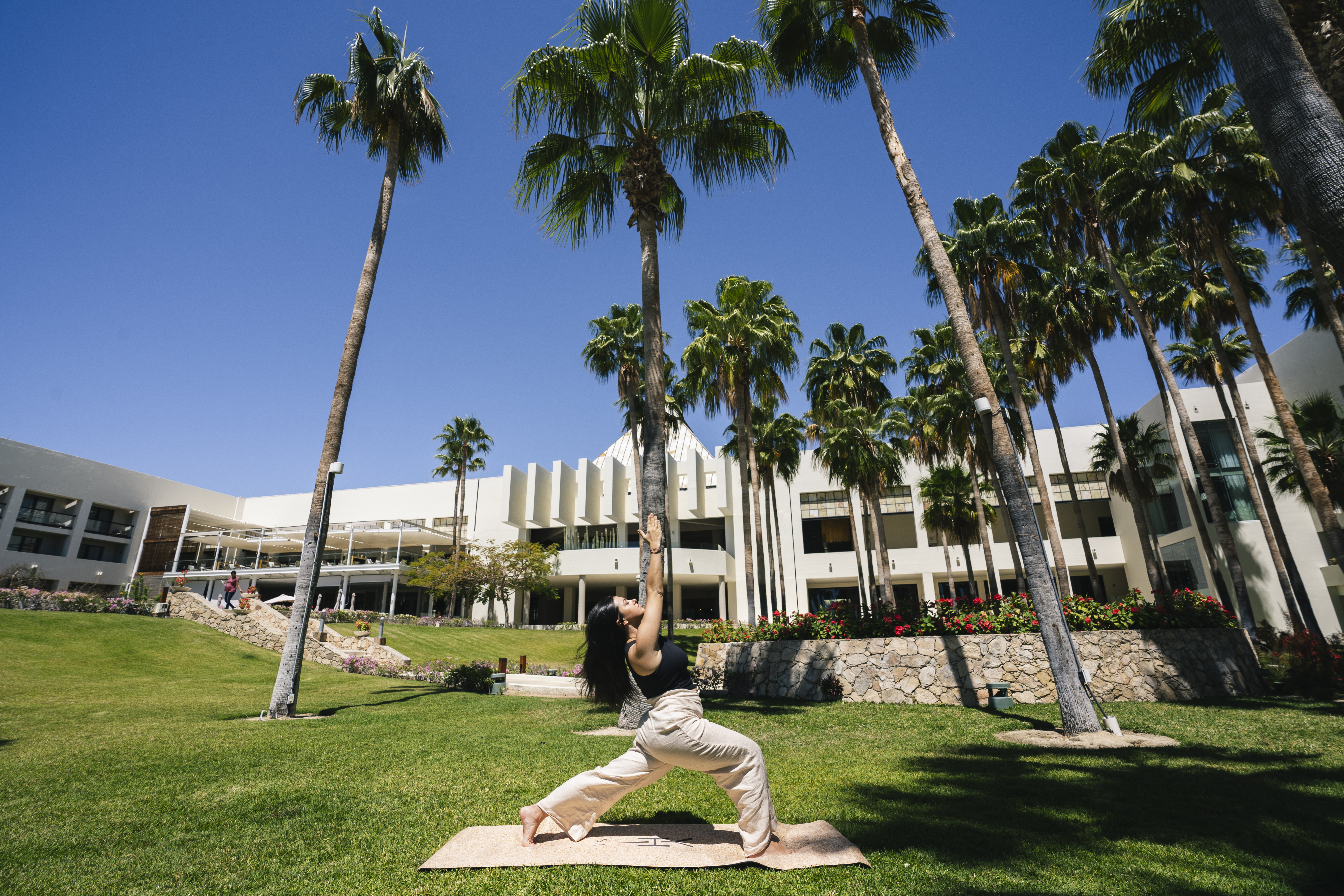 Woman in yoga pose on mat among palm trees at sunny resort.
