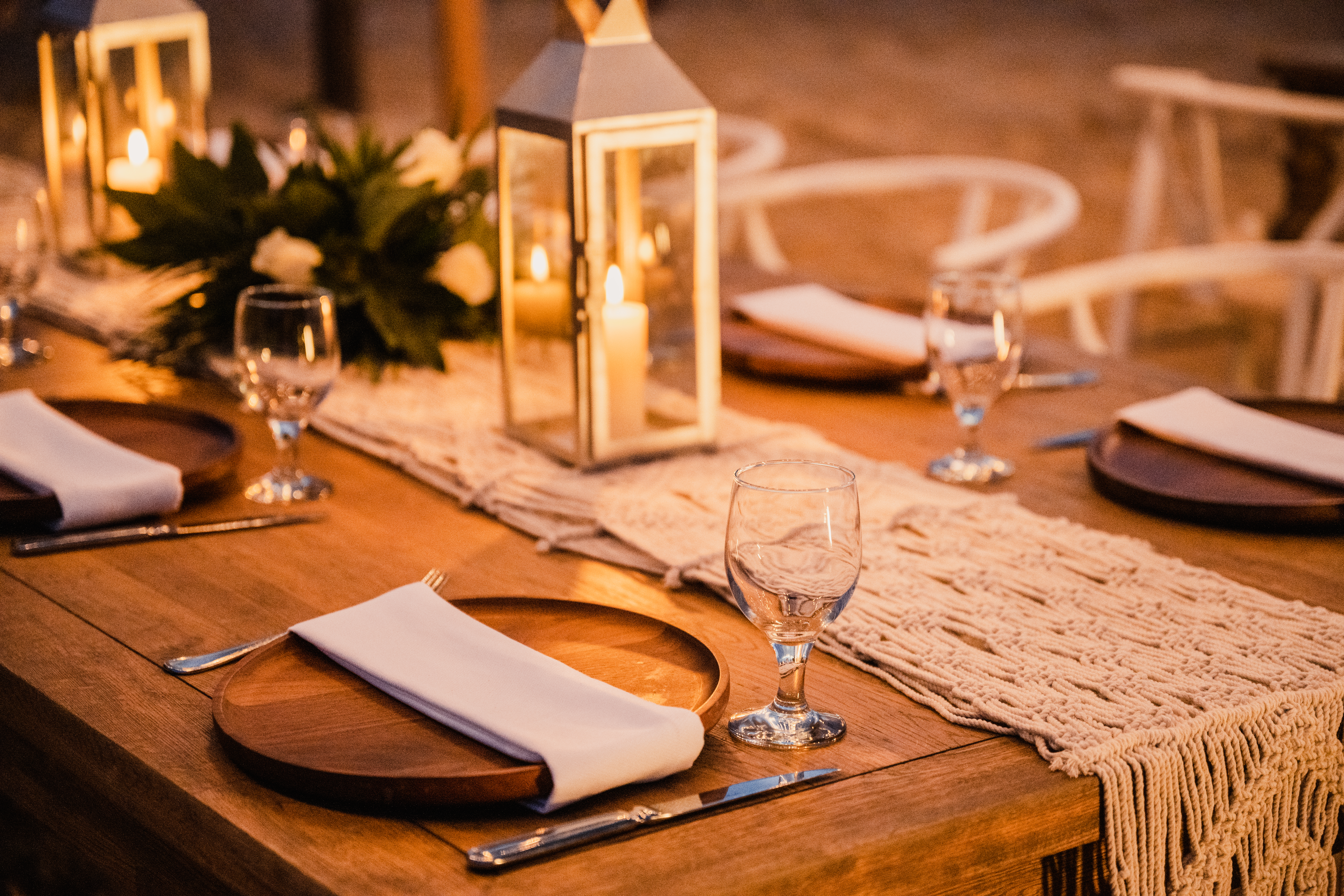 Elegant dinner table with macrame runner, candle lanterns, and white floral decor.