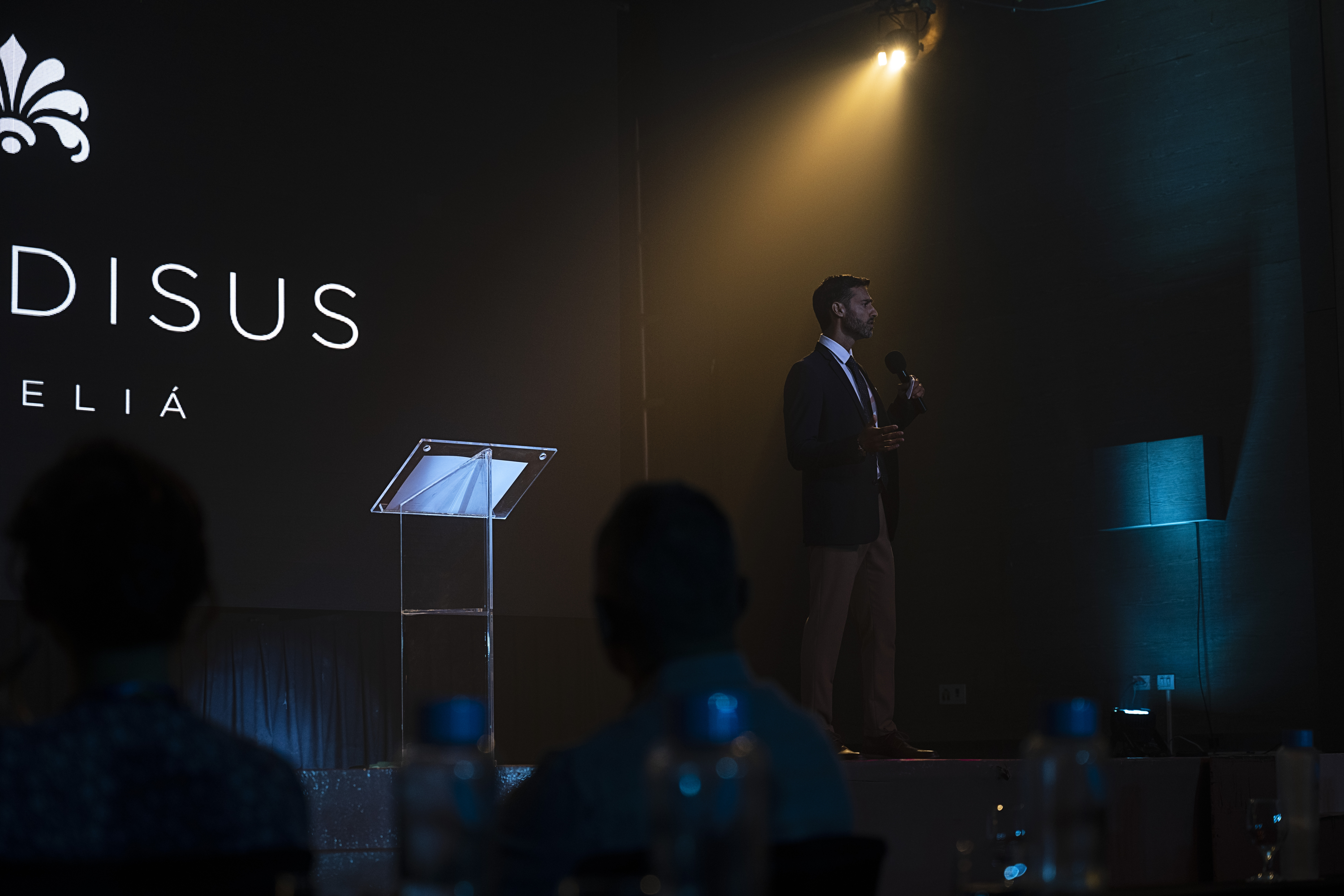 Speaker at a conference stage with microphone, DISUS ELIÁ branding.