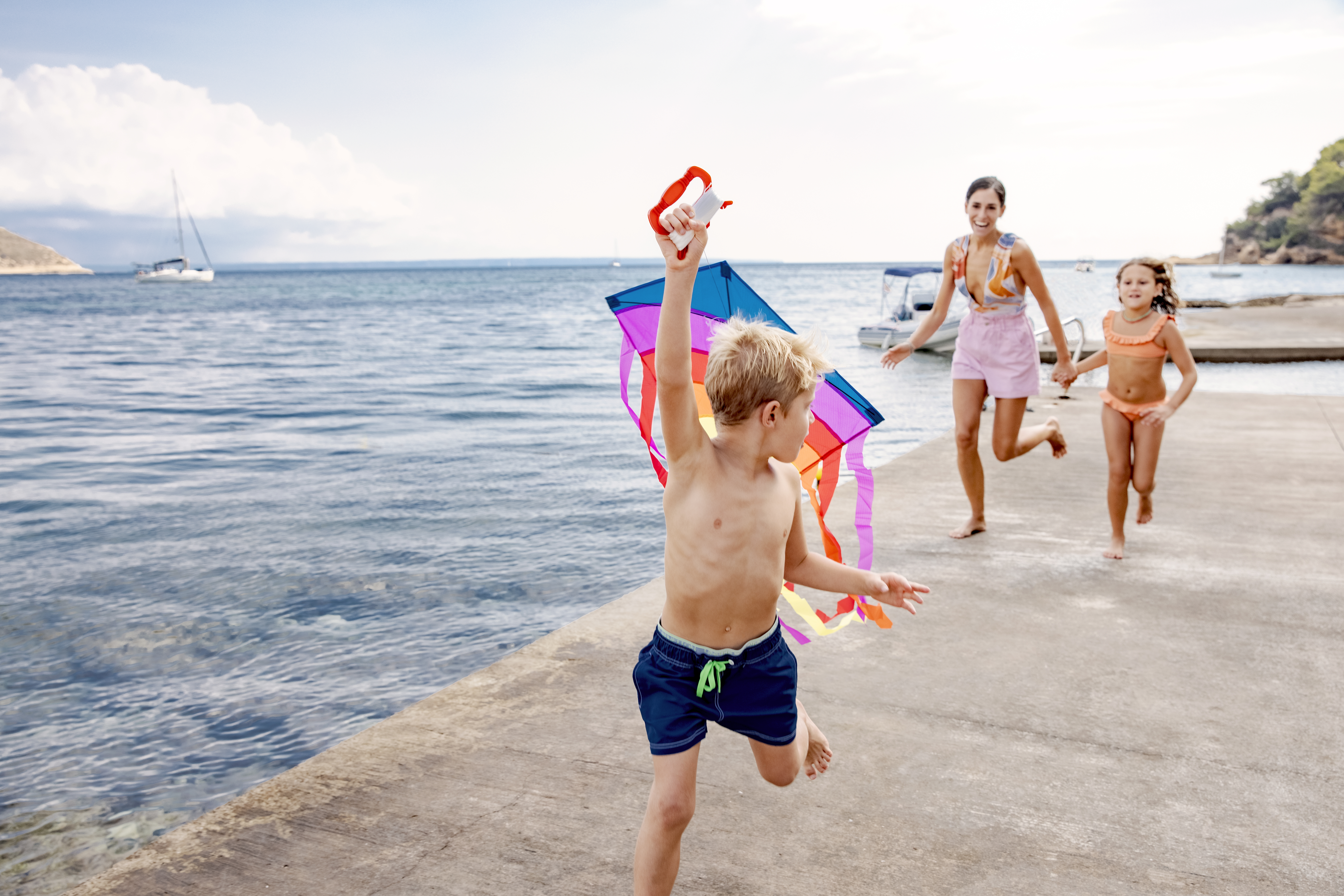 a boy running with a kite in his hand and a woman and a woman in the background