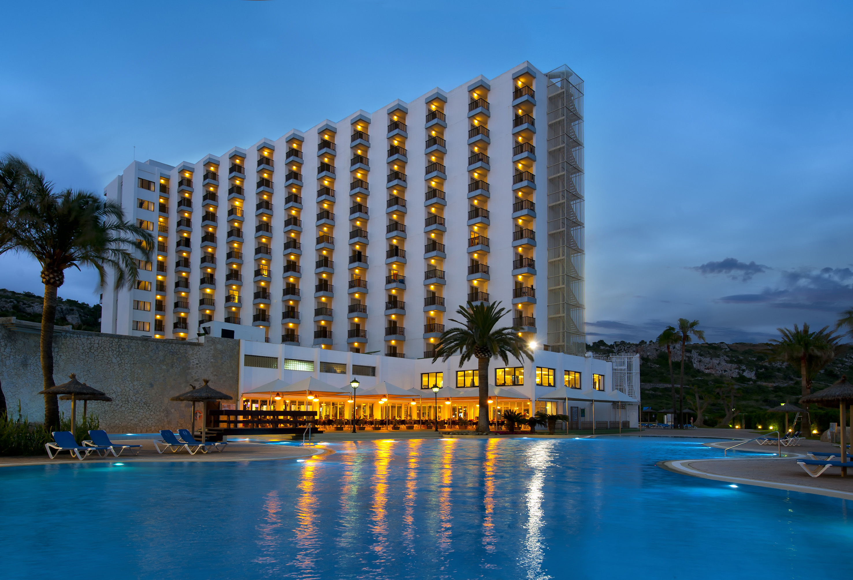 Night view of large hotel with lit balconies, swimming pool, and palm trees.