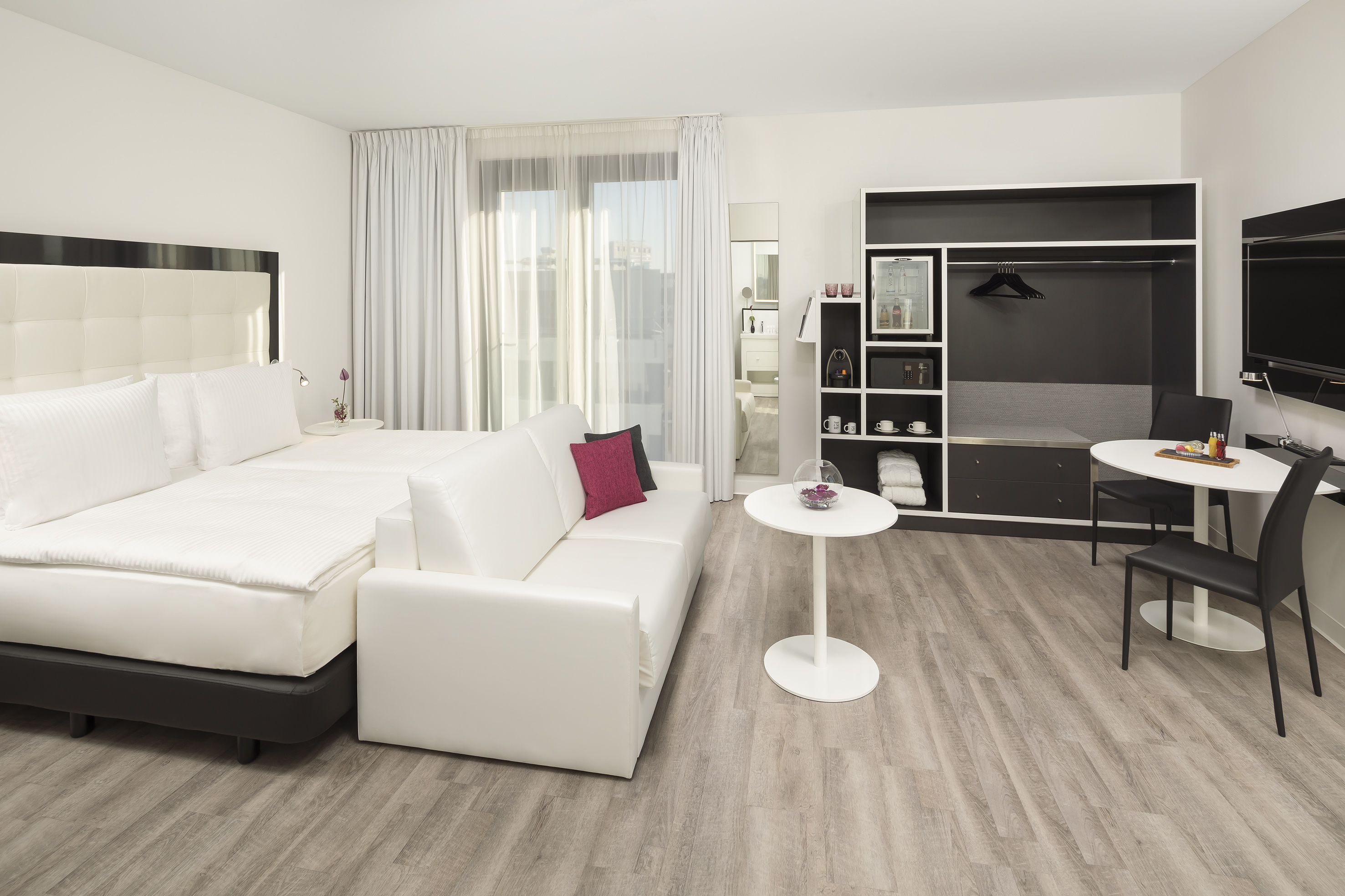 Modern hotel room with queen bed, white sofa, wood-look floor, and integrated storage.