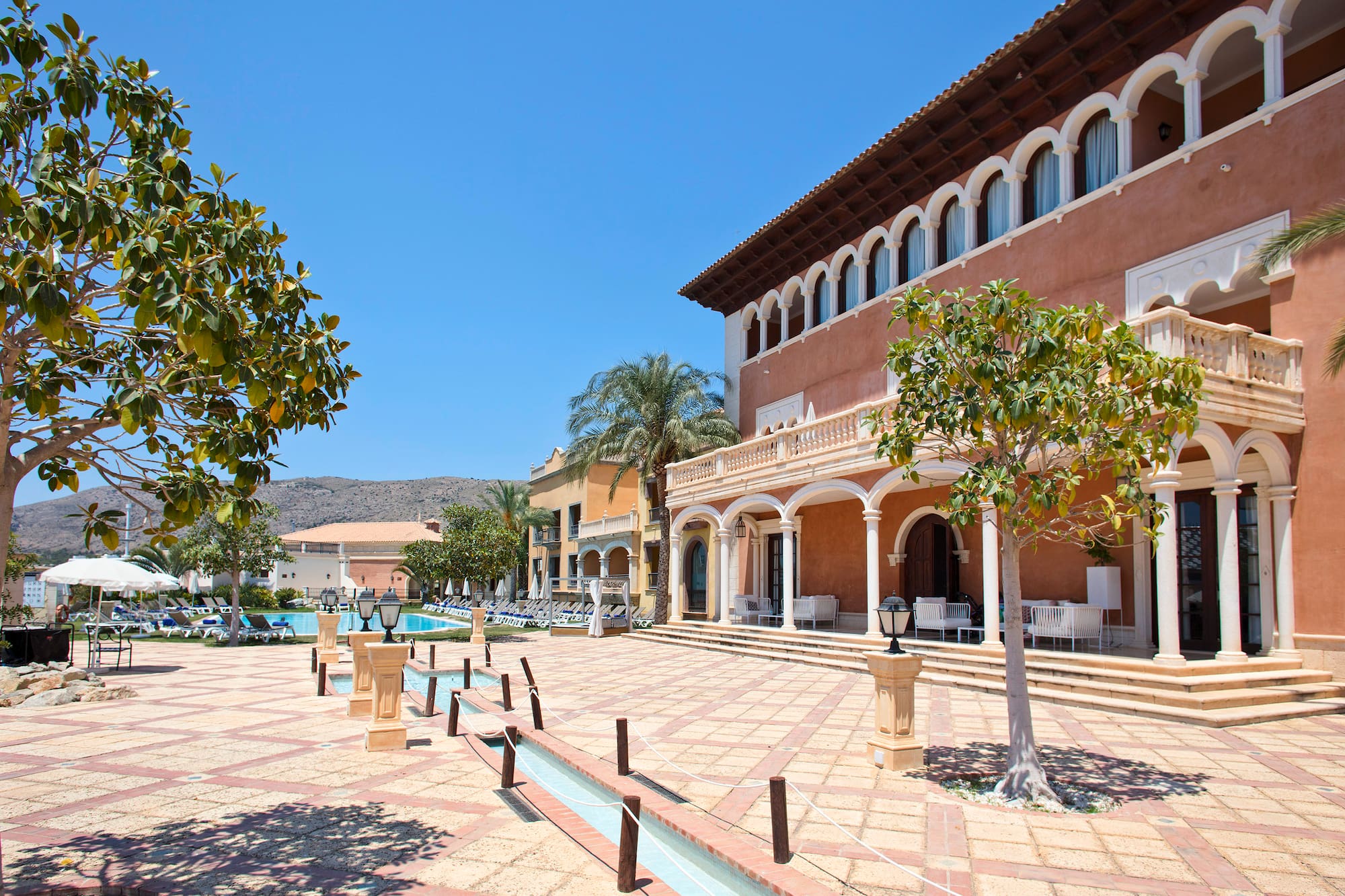 Sunny Mediterranean resort with arched buildings, swimming pool, and courtyard.