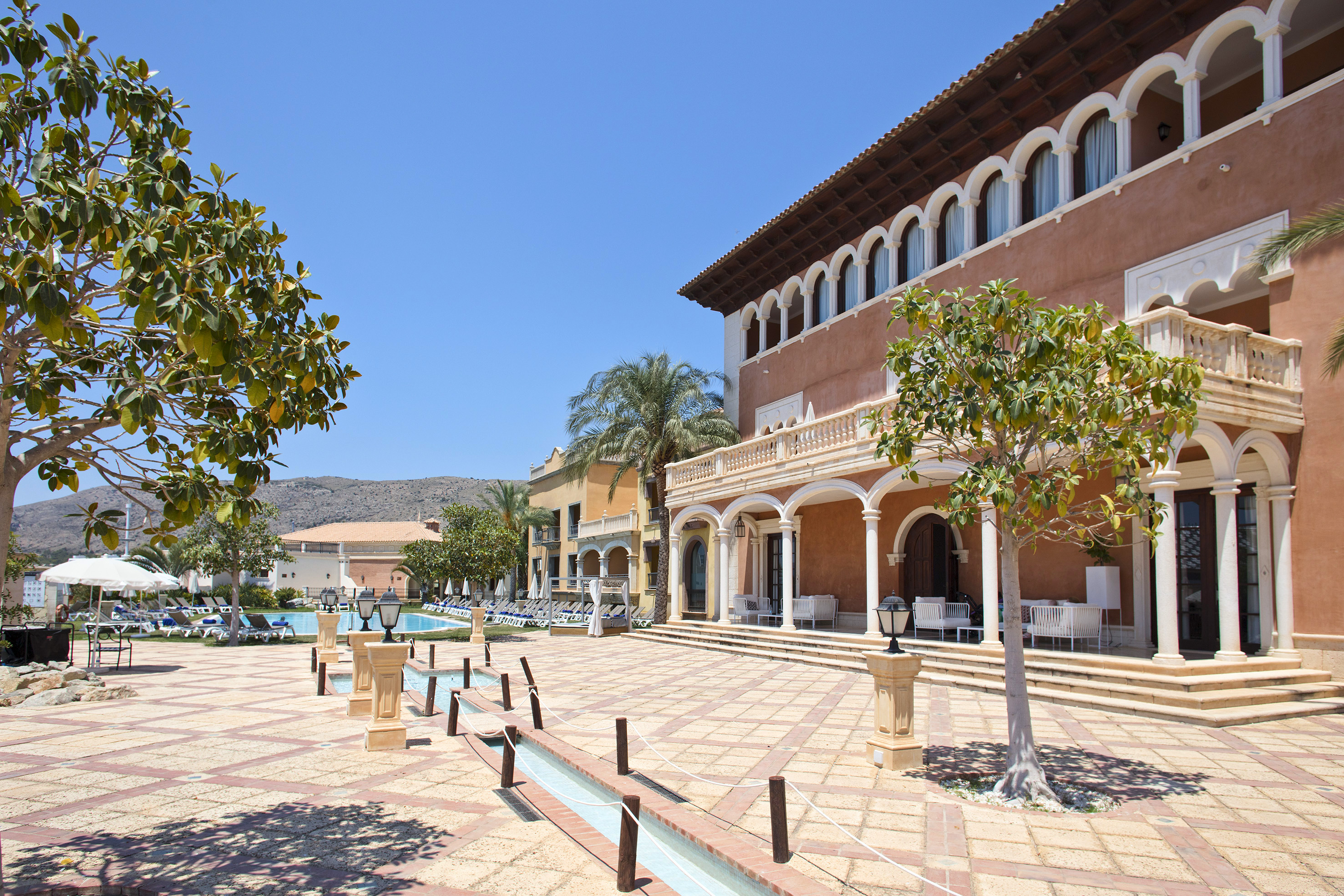 Sunny Mediterranean resort with arched buildings, swimming pool, and courtyard.