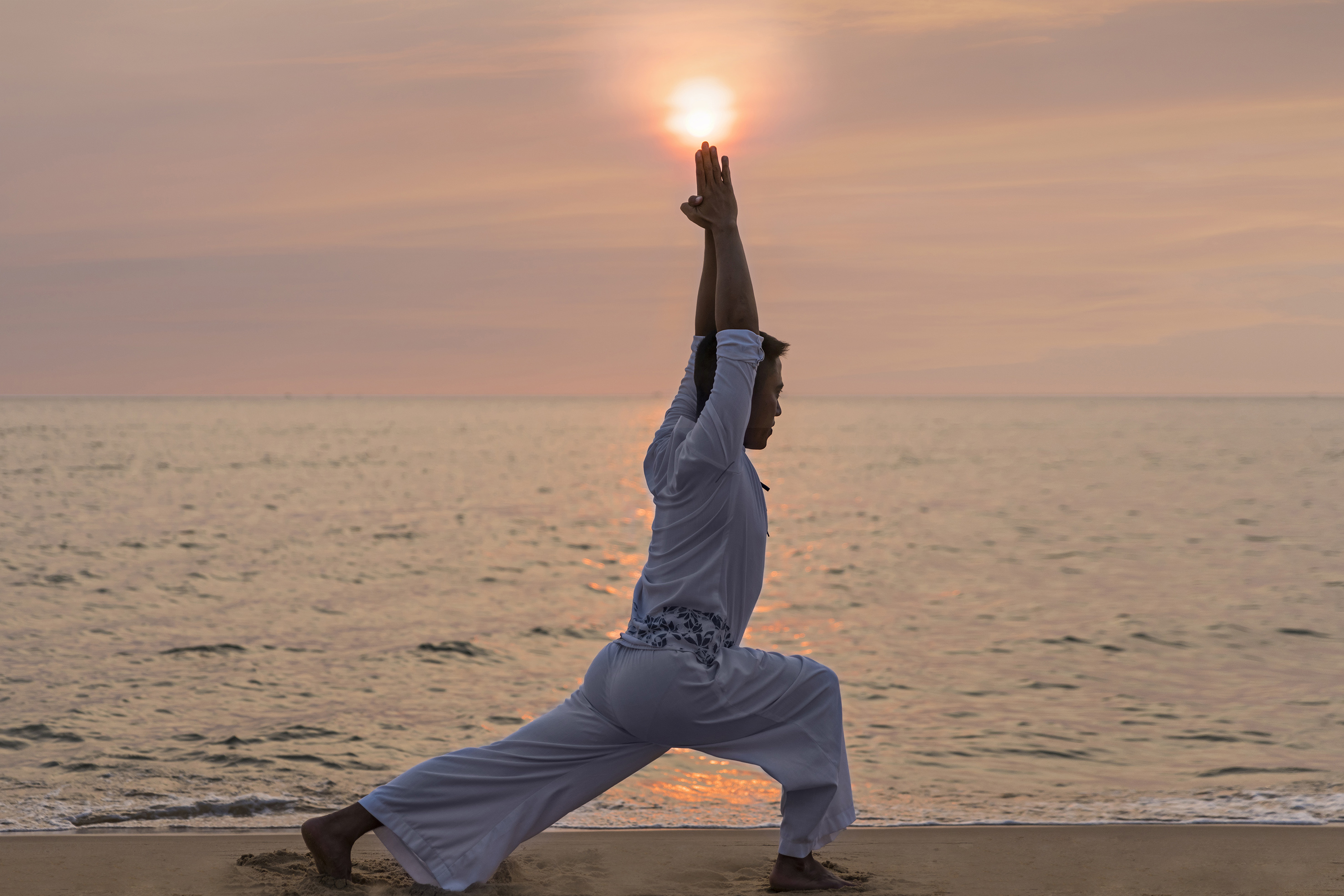 Man doing yoga Warrior I pose on beach at sunset, hands raised to sun.