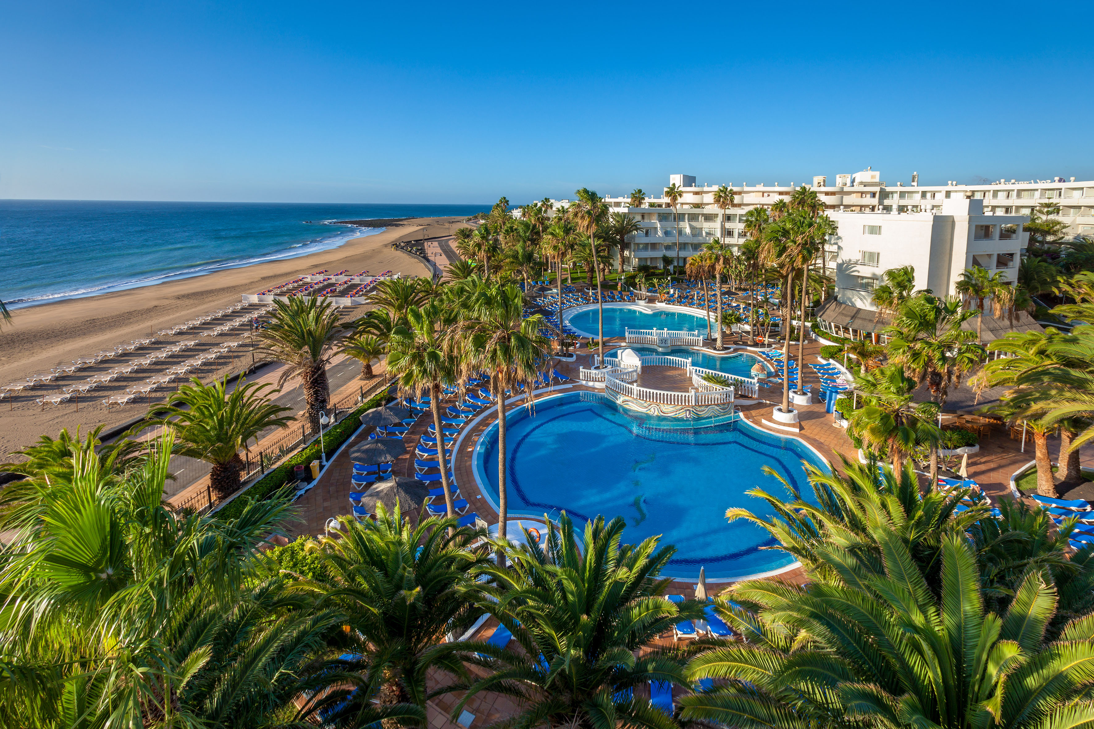 Aerial view of beachfront resort, multiple pools, palm trees, and sandy beach.
