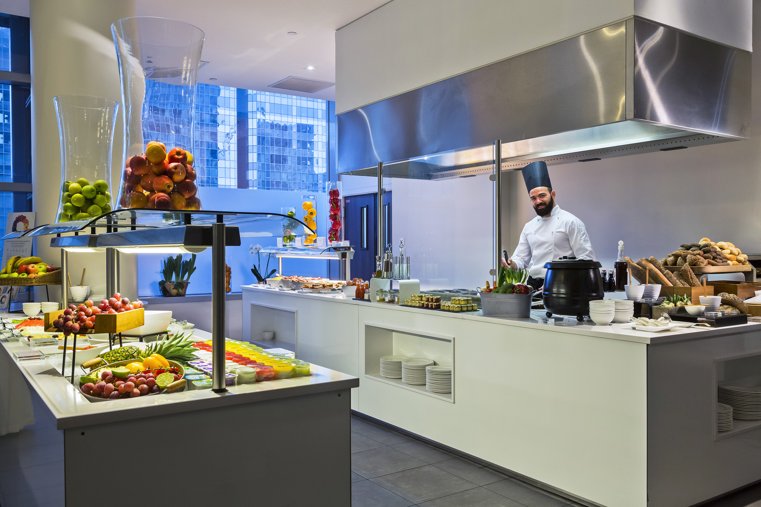 Hotel breakfast buffet with chef, fresh fruits, bread, and modern food displays.