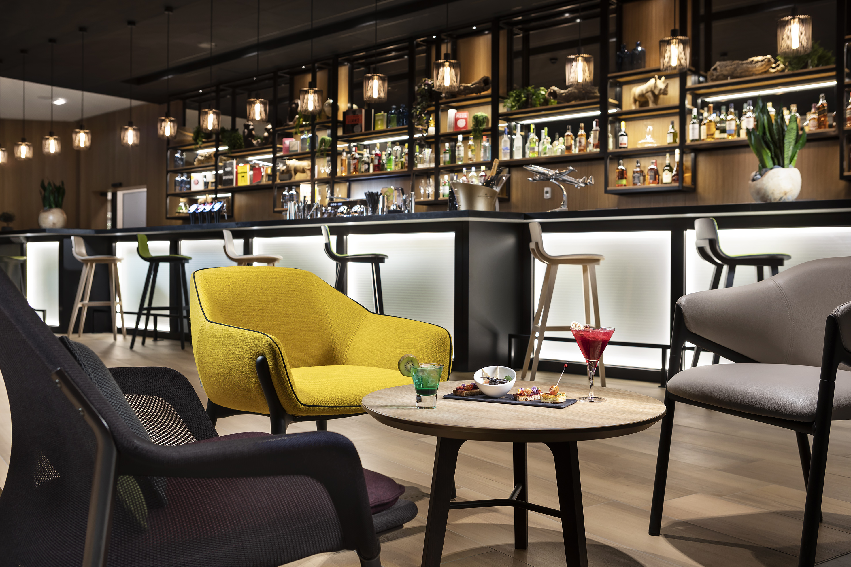 Modern lounge bar with yellow armchair, cocktails, and lit bottle display.