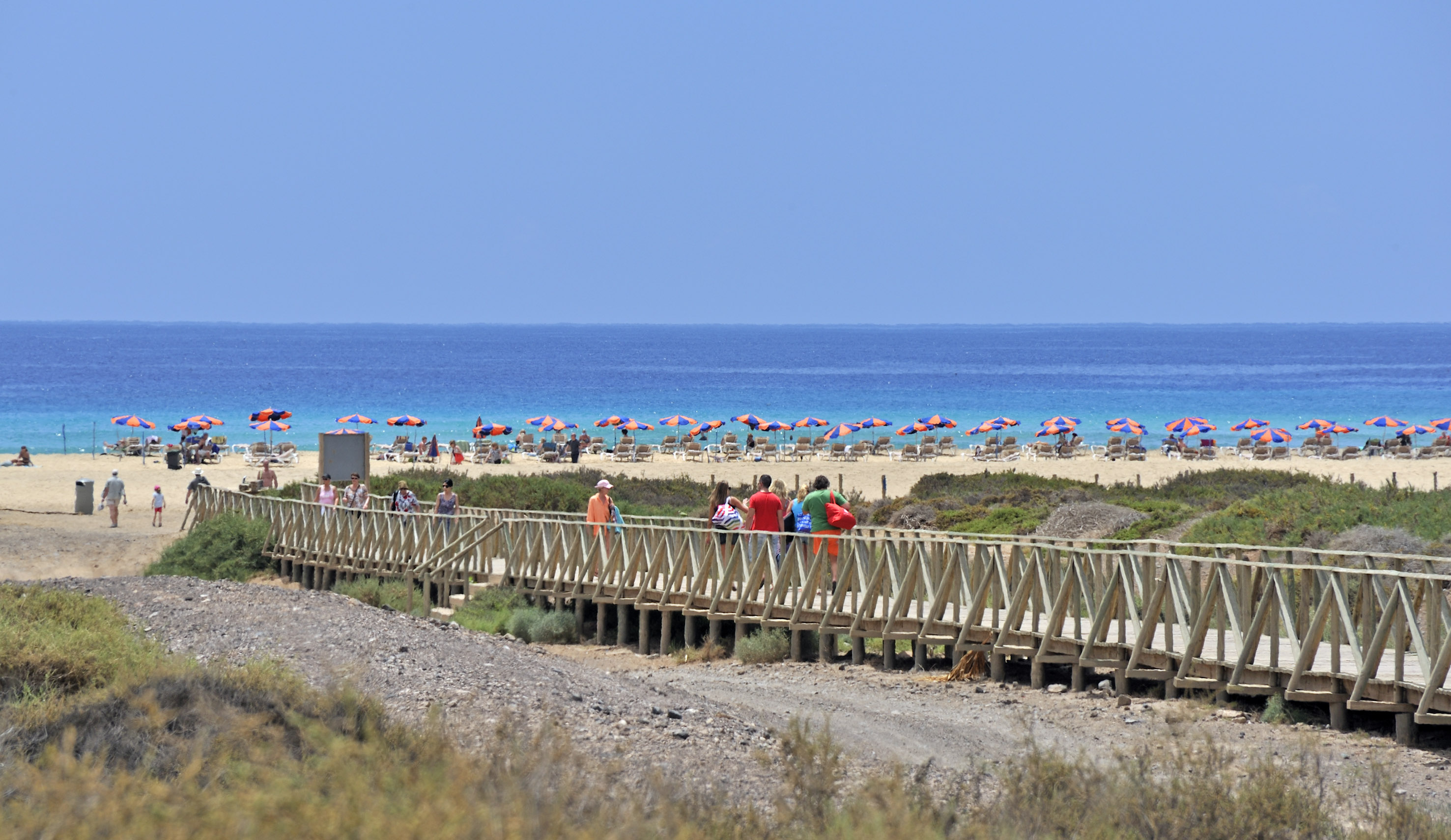 People on boardwalk to sunny beach, orange umbrellas, clear blue ocean.
