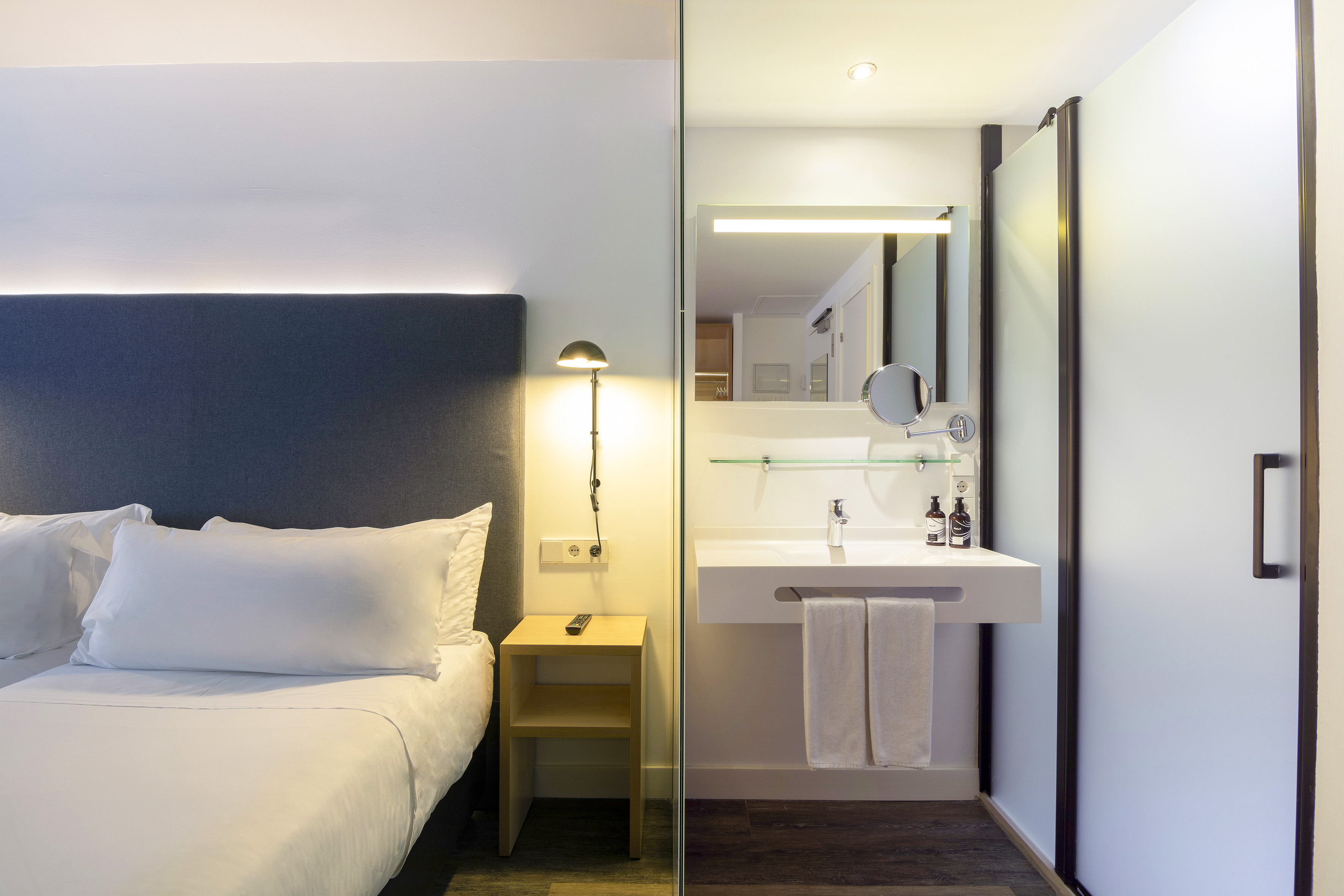 Modern hotel room with bed, side table, and open bathroom vanity and shower.