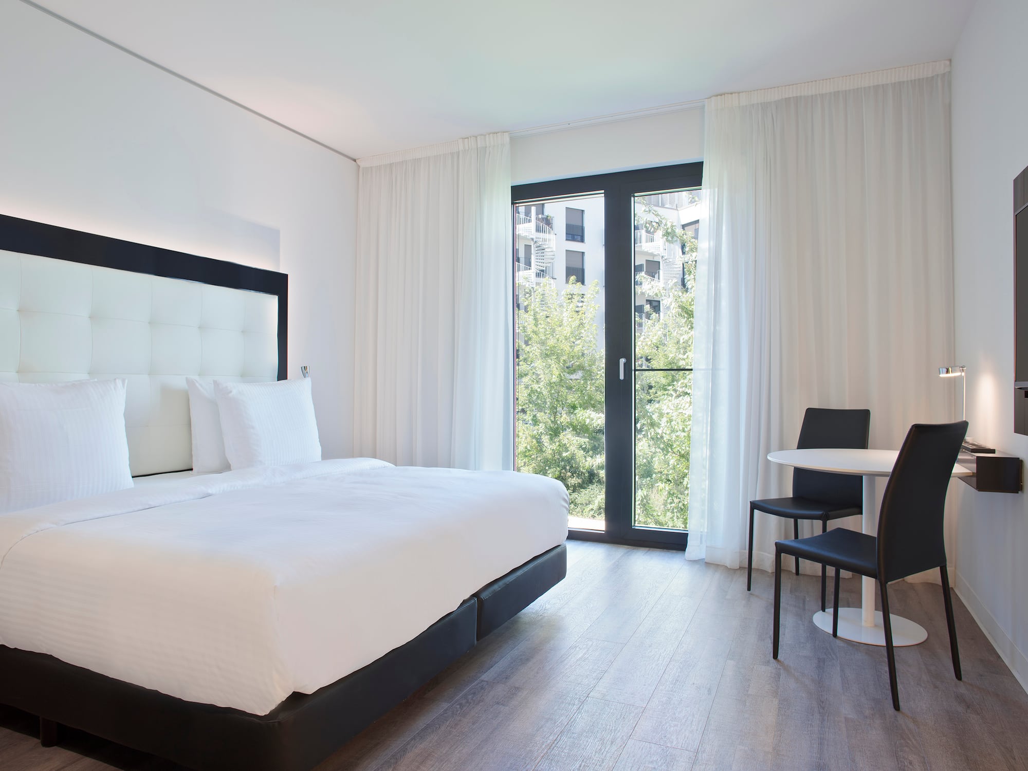 Minimalist hotel room: white bed, large window with trees, table, two chairs.
