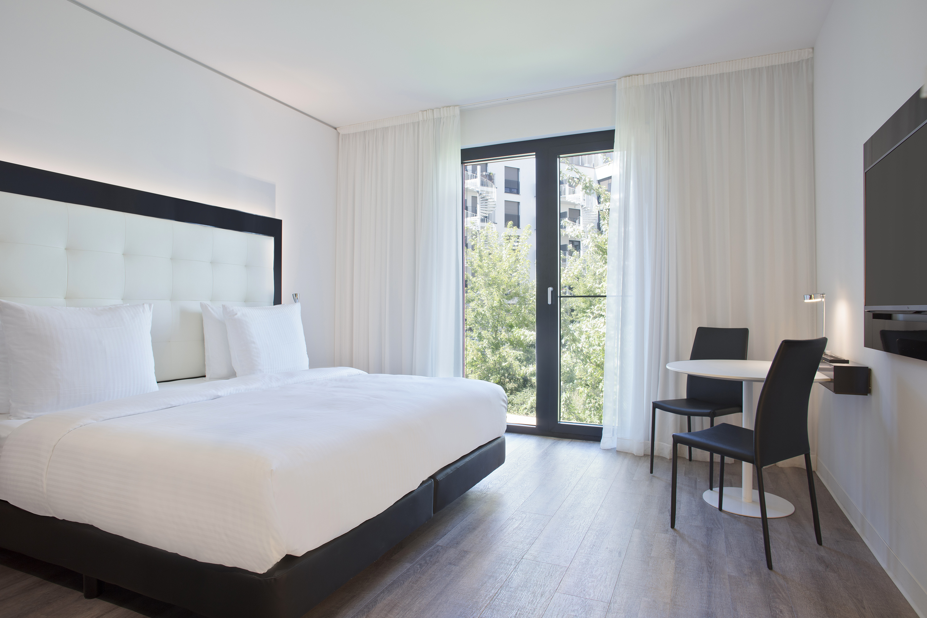 Minimalist hotel room: white bed, large window with trees, table, two chairs.