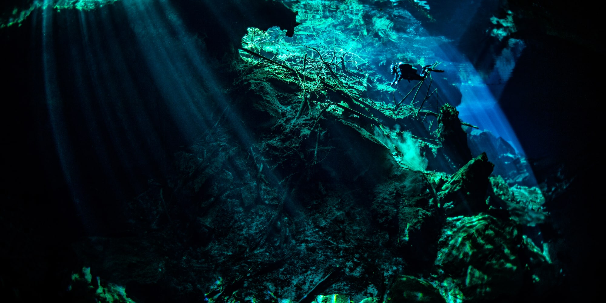 Diver in a cenote, sunlight beaming through clear blue water into the cavern.