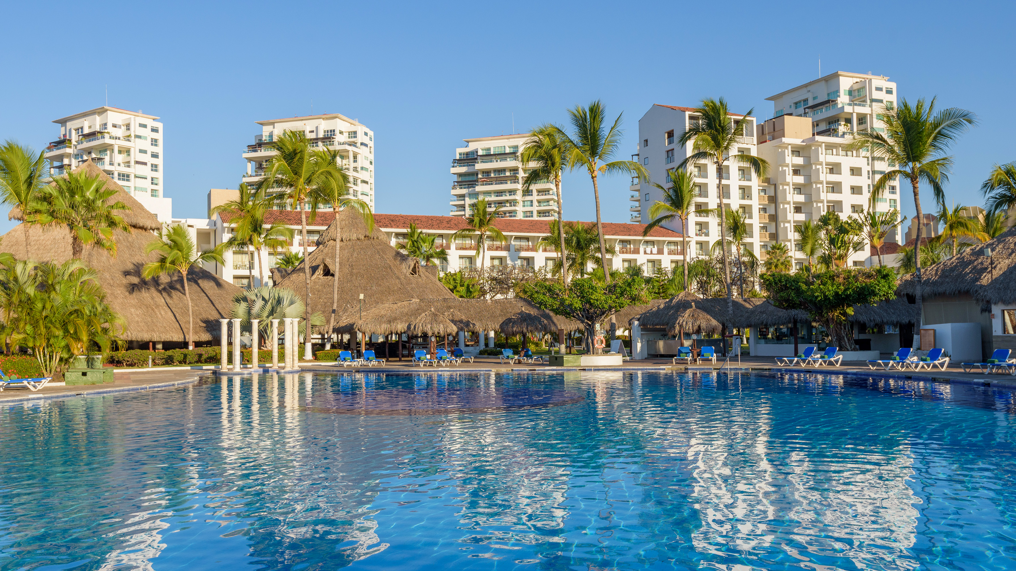 Tropical resort pool, palm trees, palapas, and modern hotel buildings.