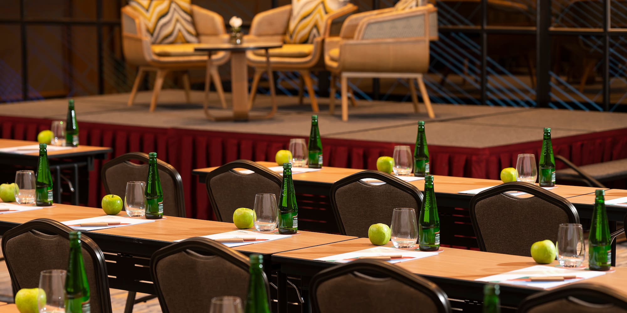 a room with tables and chairs and a stage with chairs and a stage with chairs and a table with green bottles and apples