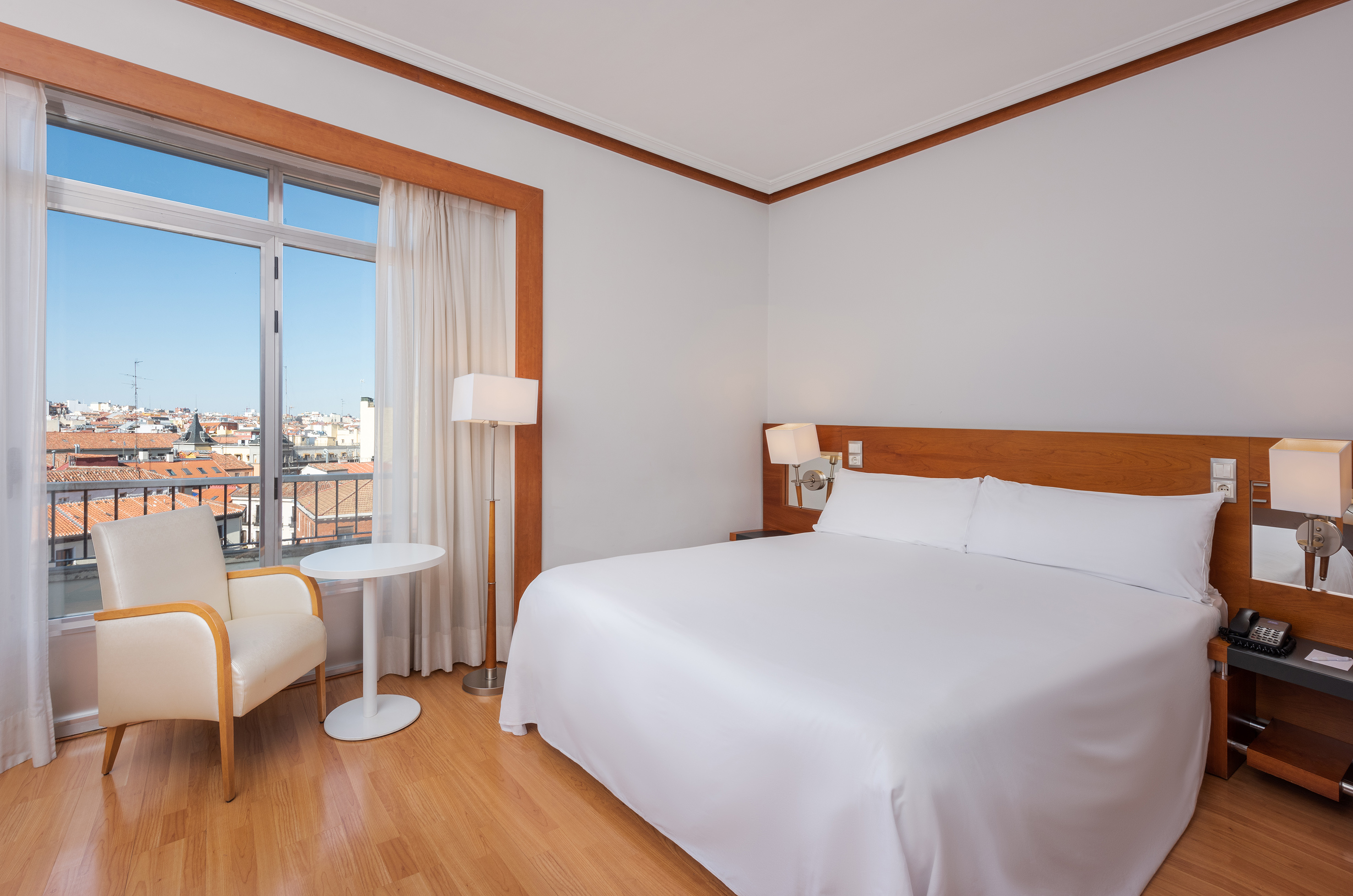 Bright hotel room with white king bed, city view, armchair, and wooden floor.