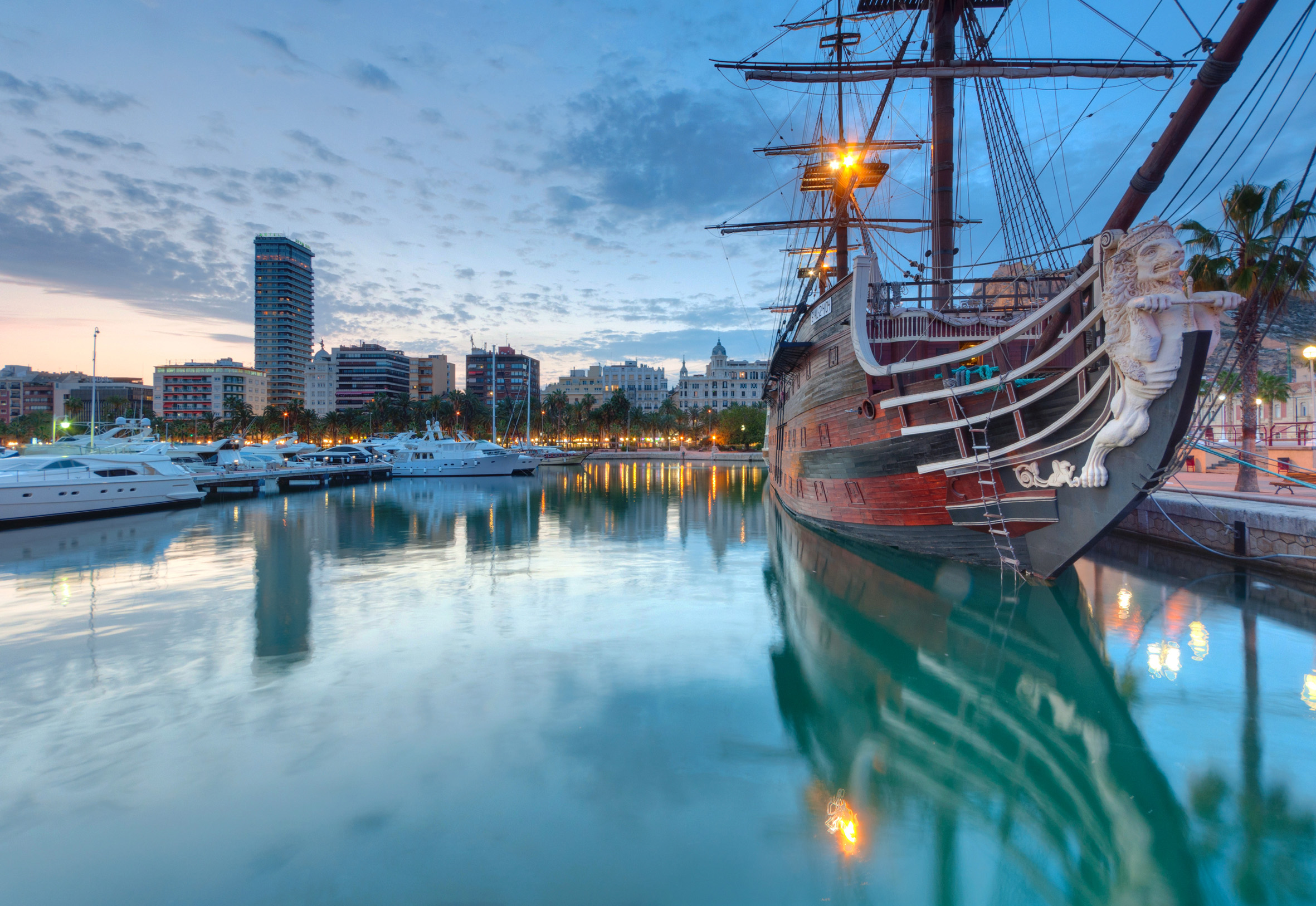 Historic sailing ship with lion figurehead and modern yachts in a twilight marina.