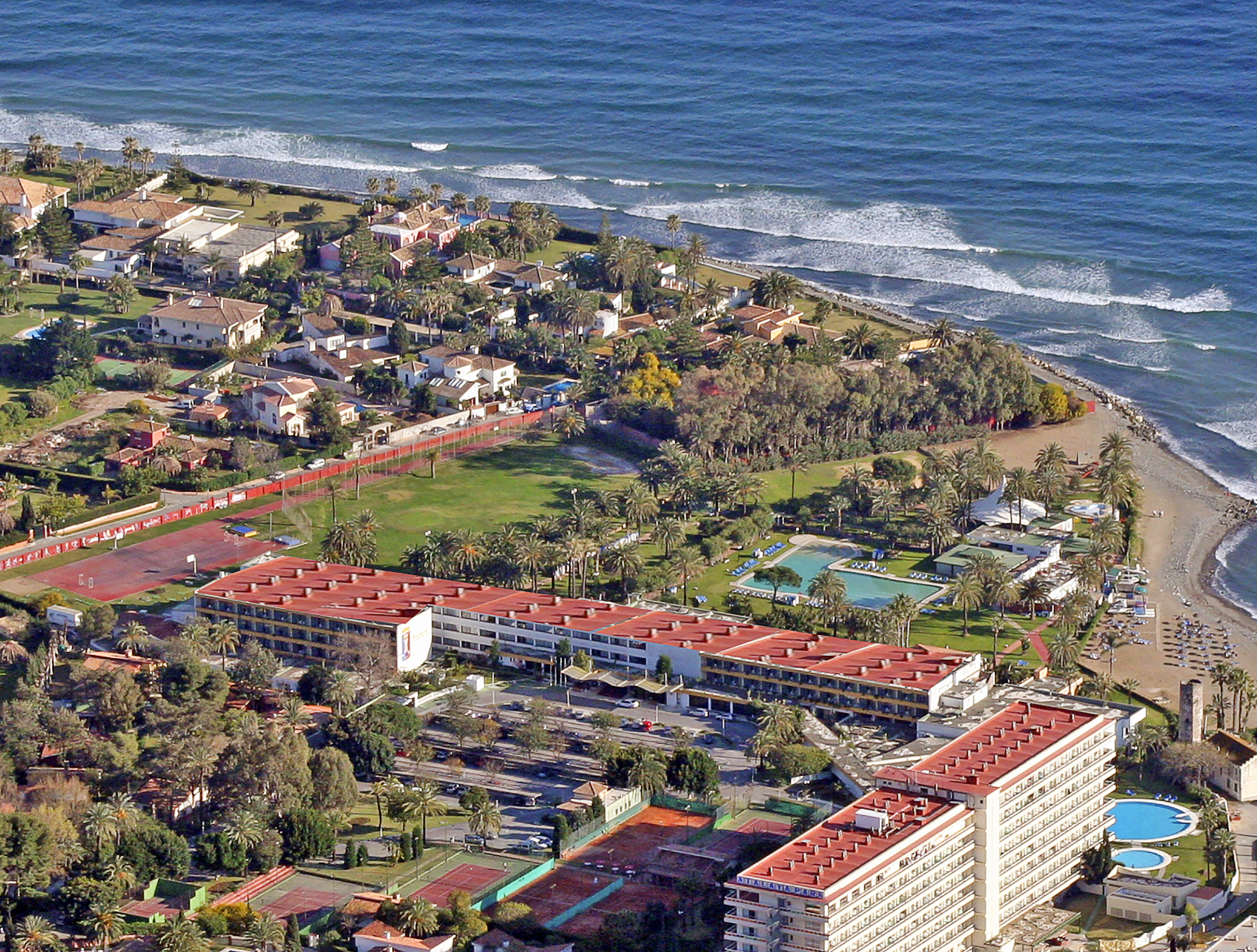 Aerial view: beachfront resort with hotels, swimming pools, tennis courts, palm trees.