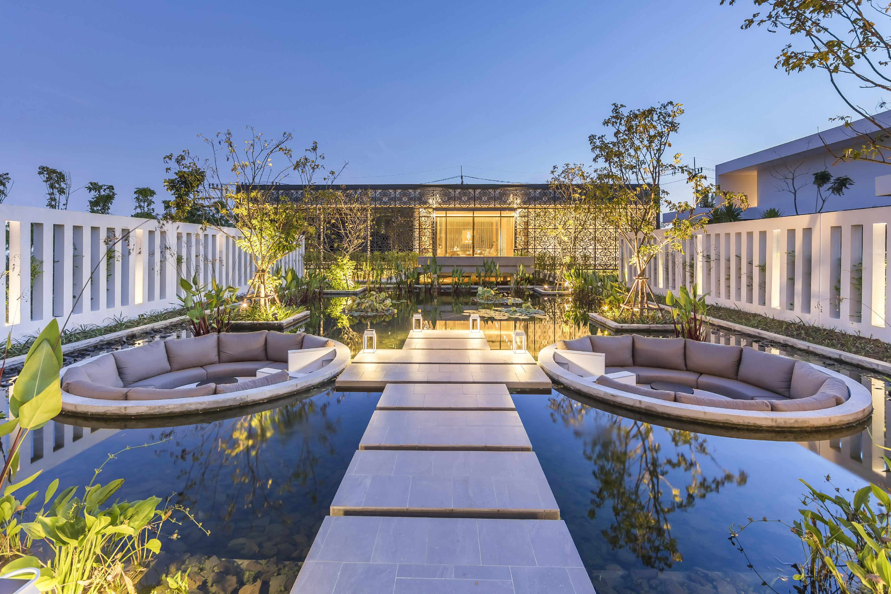 Illuminated modern outdoor lounge with pond, stepping path, and elegant building at dusk.