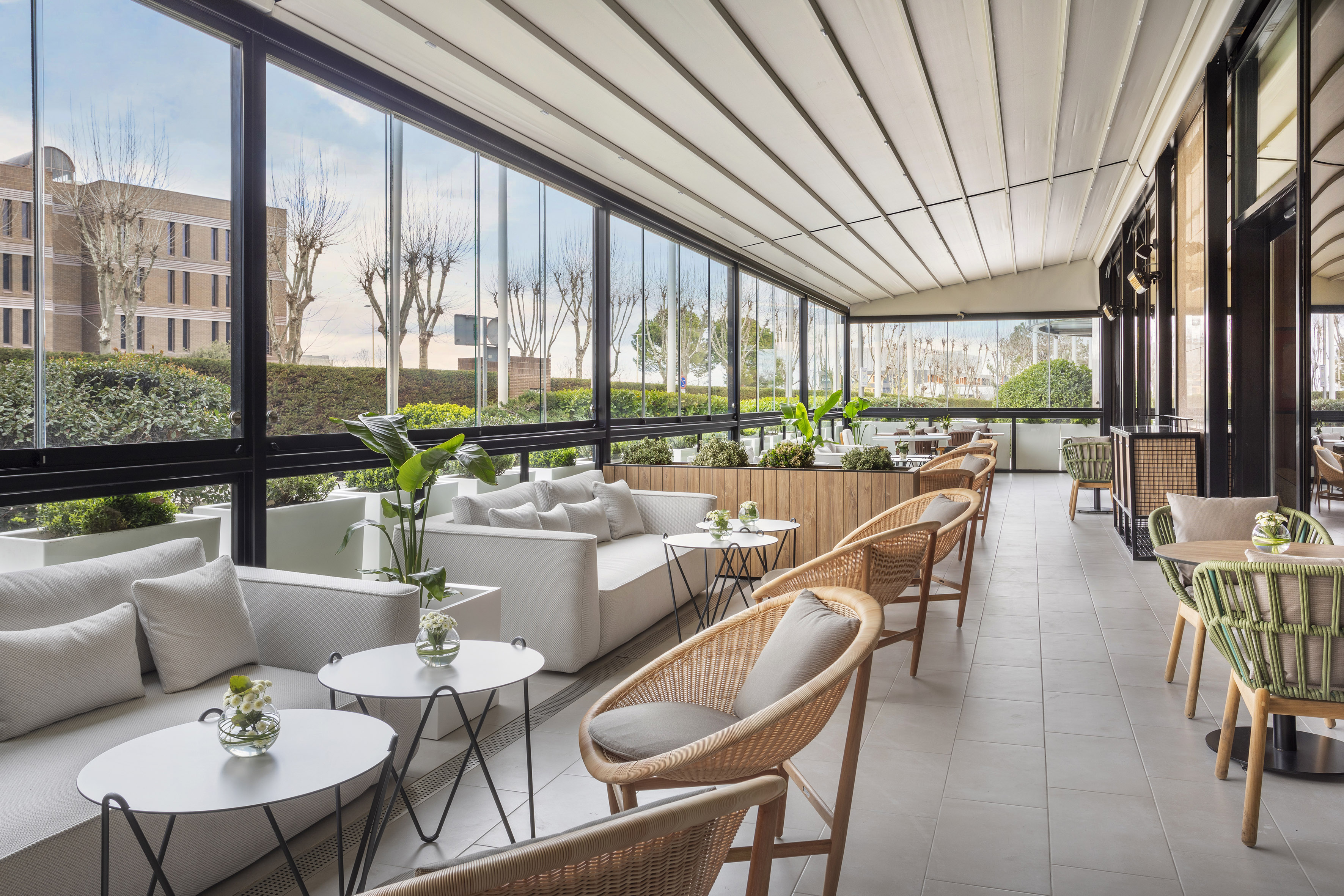 Modern patio lounge with glass walls, retractable roof, sofas, and wicker chairs.