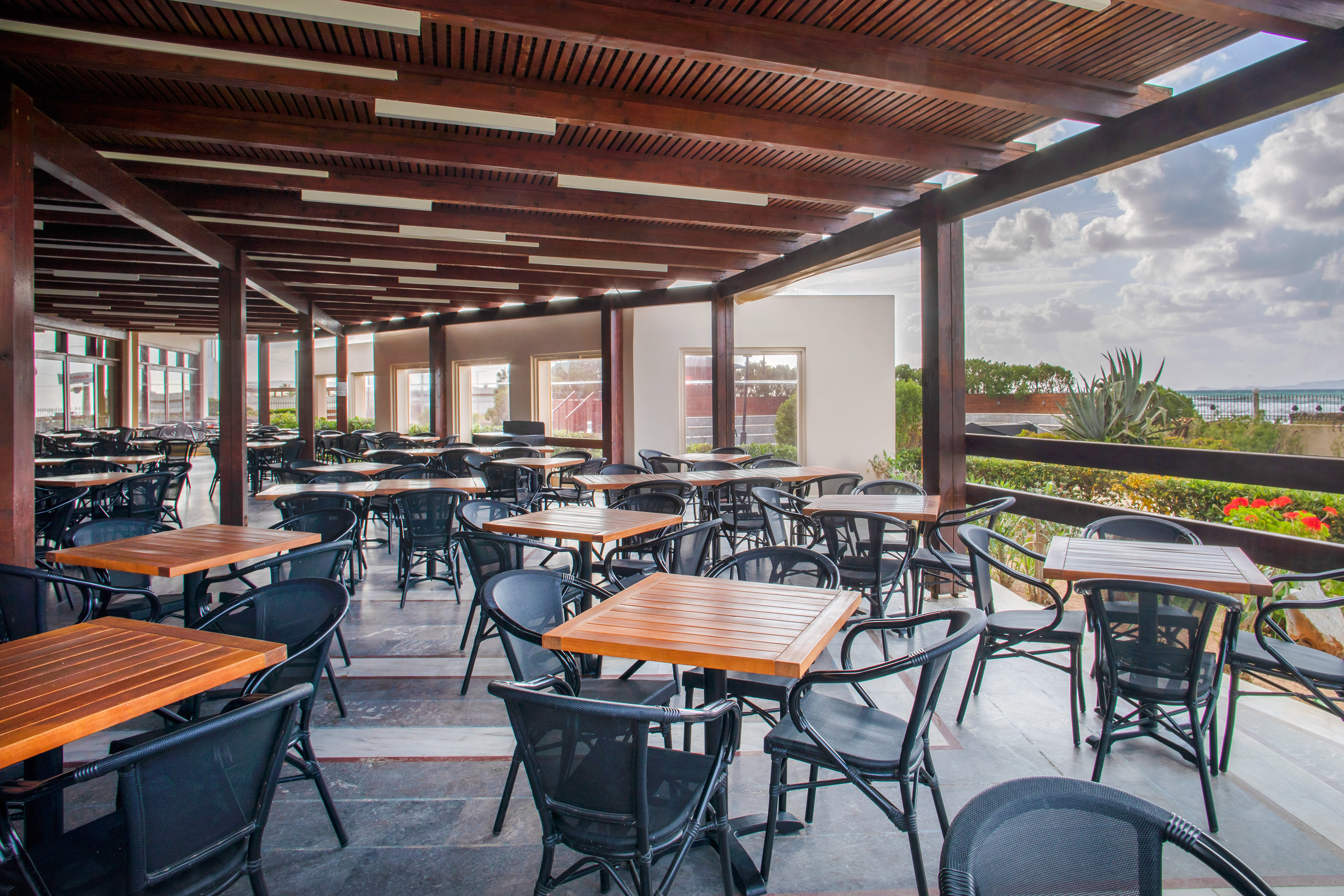 Resort outdoor dining patio with wooden tables, black chairs, and ocean view.