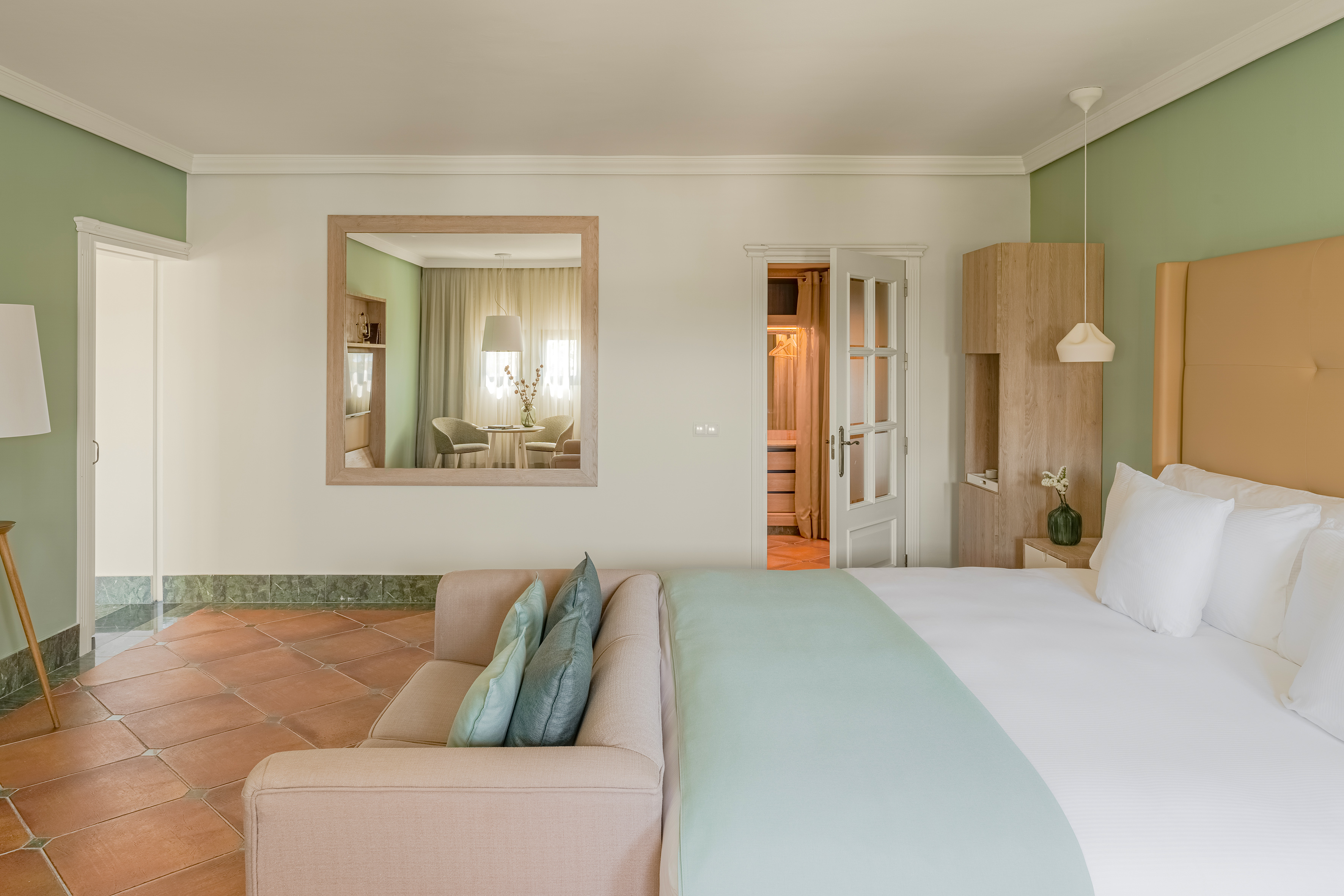 Hotel suite with bed, sofa, terracotta floor, green wall, and large mirror.