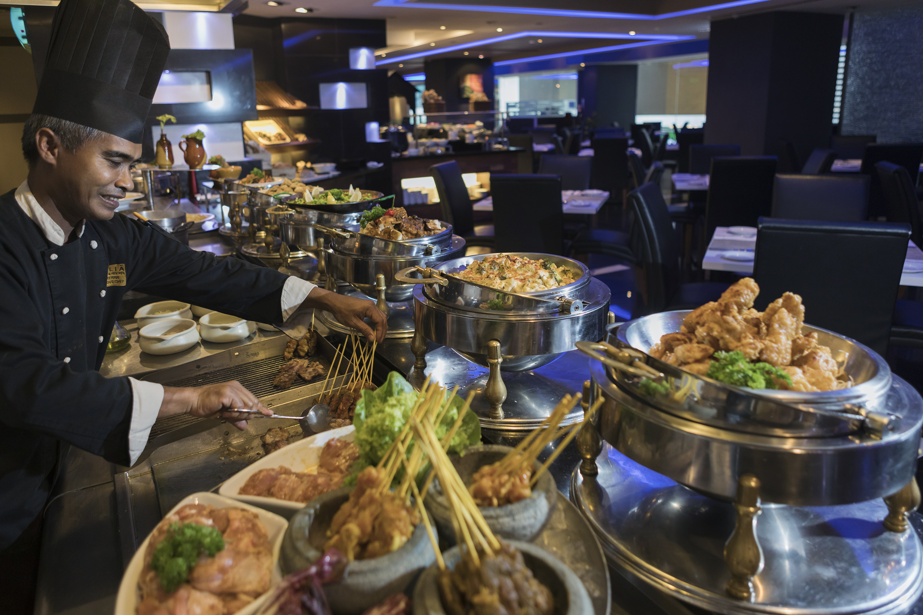 Smiling chef serving satay and hot dishes at a hotel restaurant buffet.