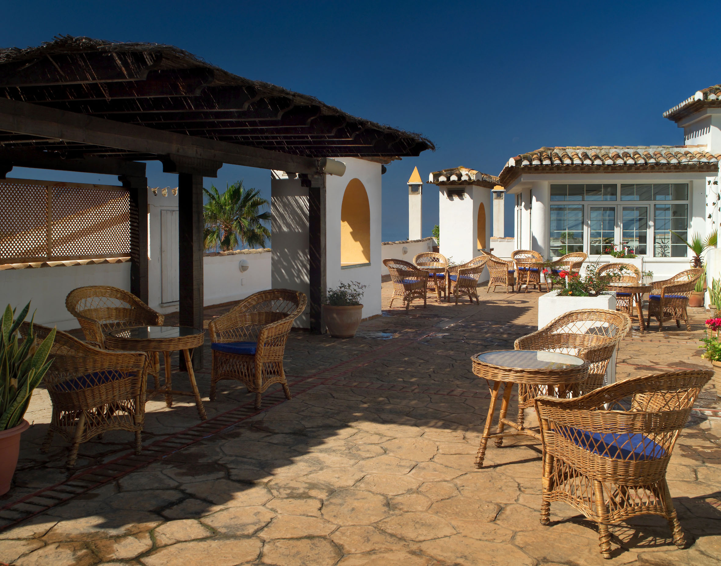 Mediterranean terrace with wicker chairs, tables, pergola, sunny blue sky.