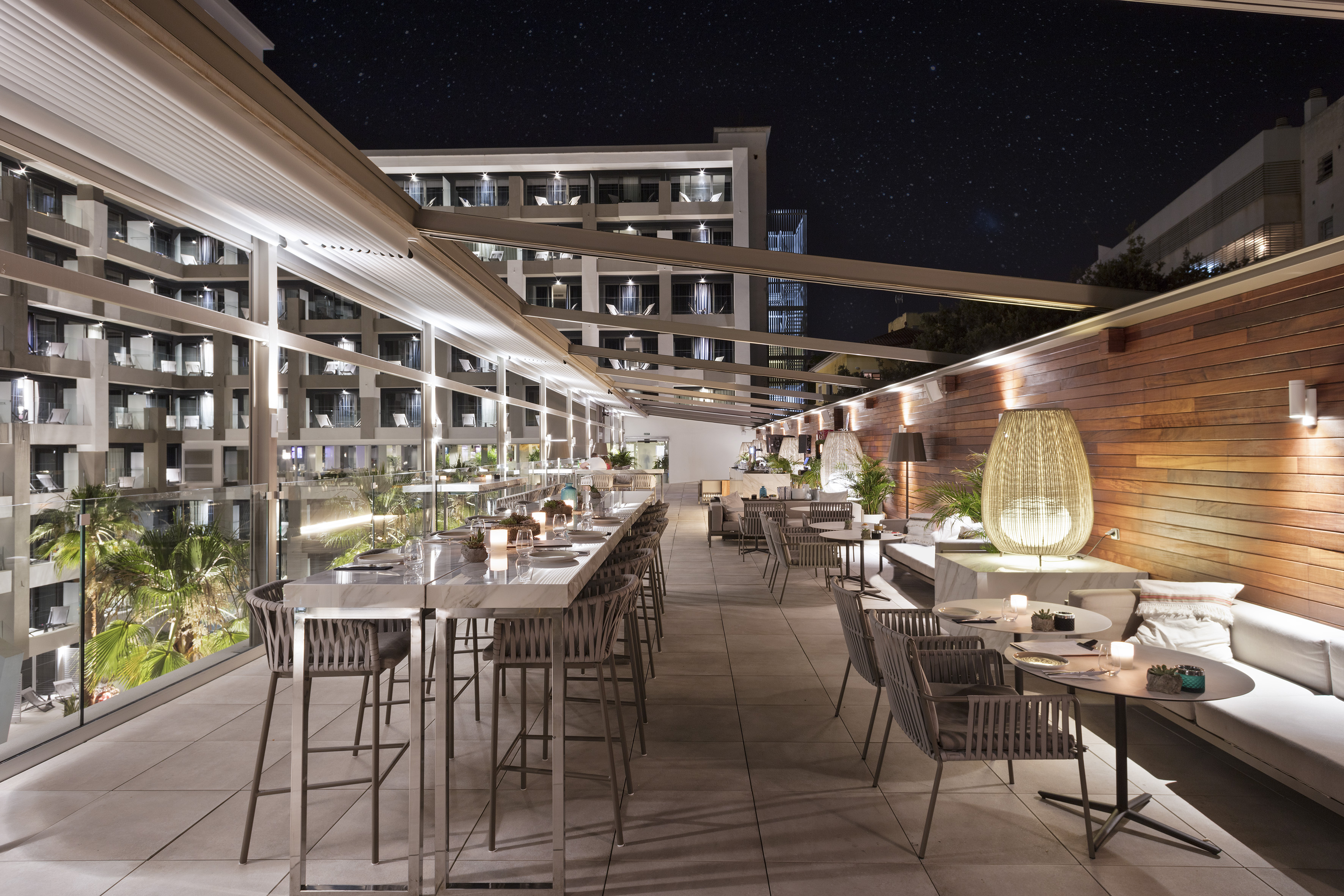 Modern rooftop bar at night with outdoor tables, hotel building, and starry sky.