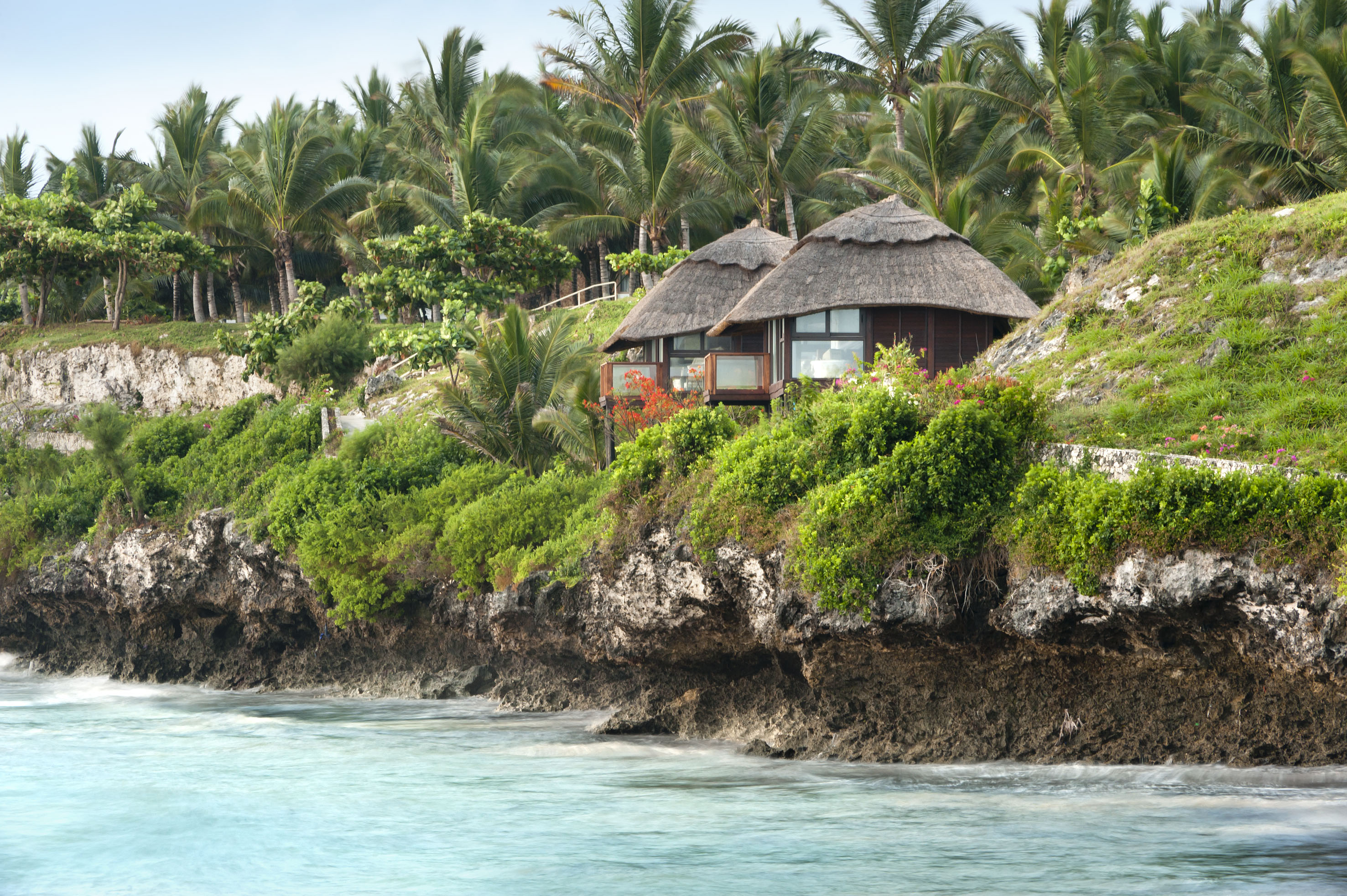 Thatched bungalows on tropical cliff above turquoise ocean with palm trees.