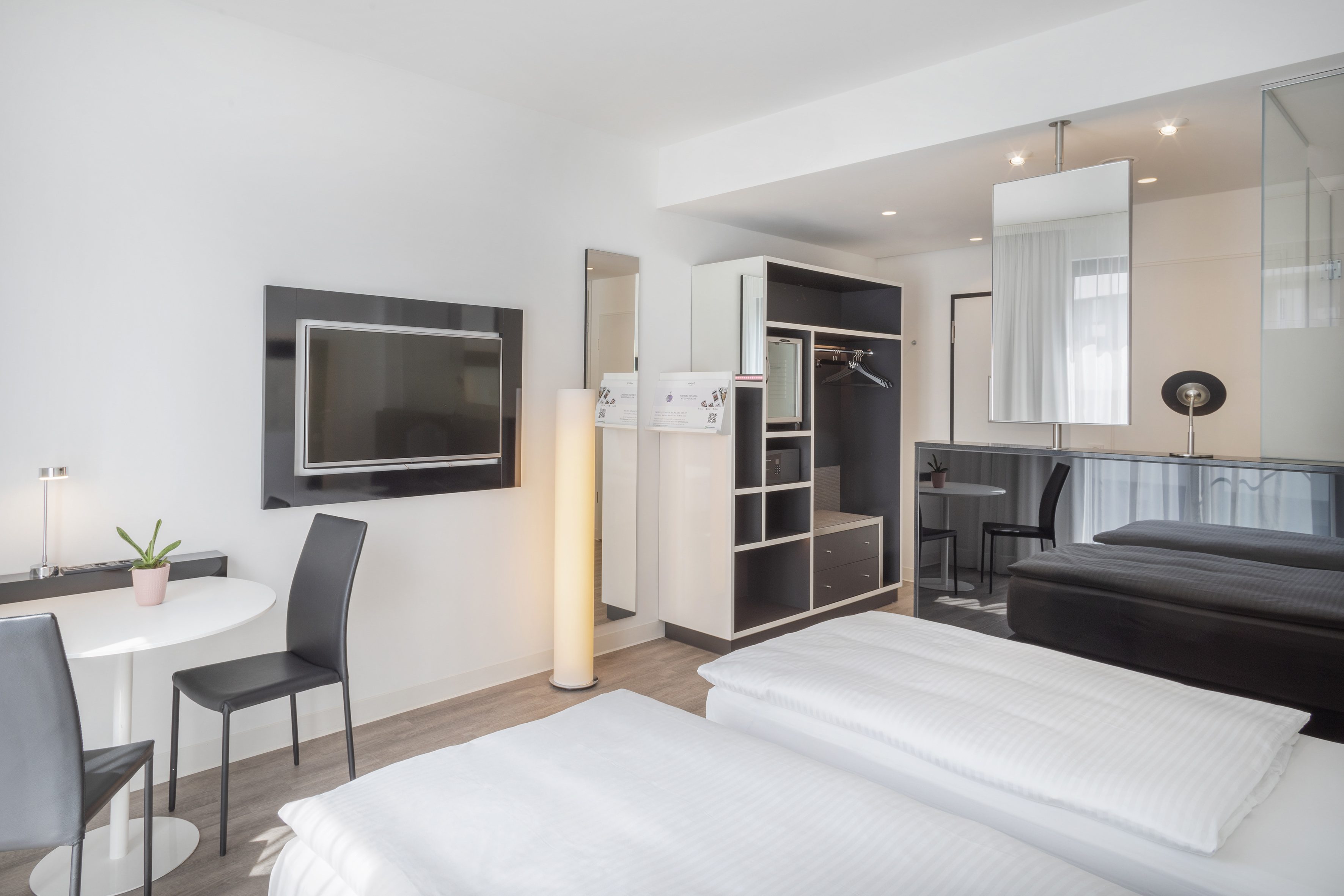 Minimalist modern hotel room with two beds, wall TV, table, and open wardrobe.